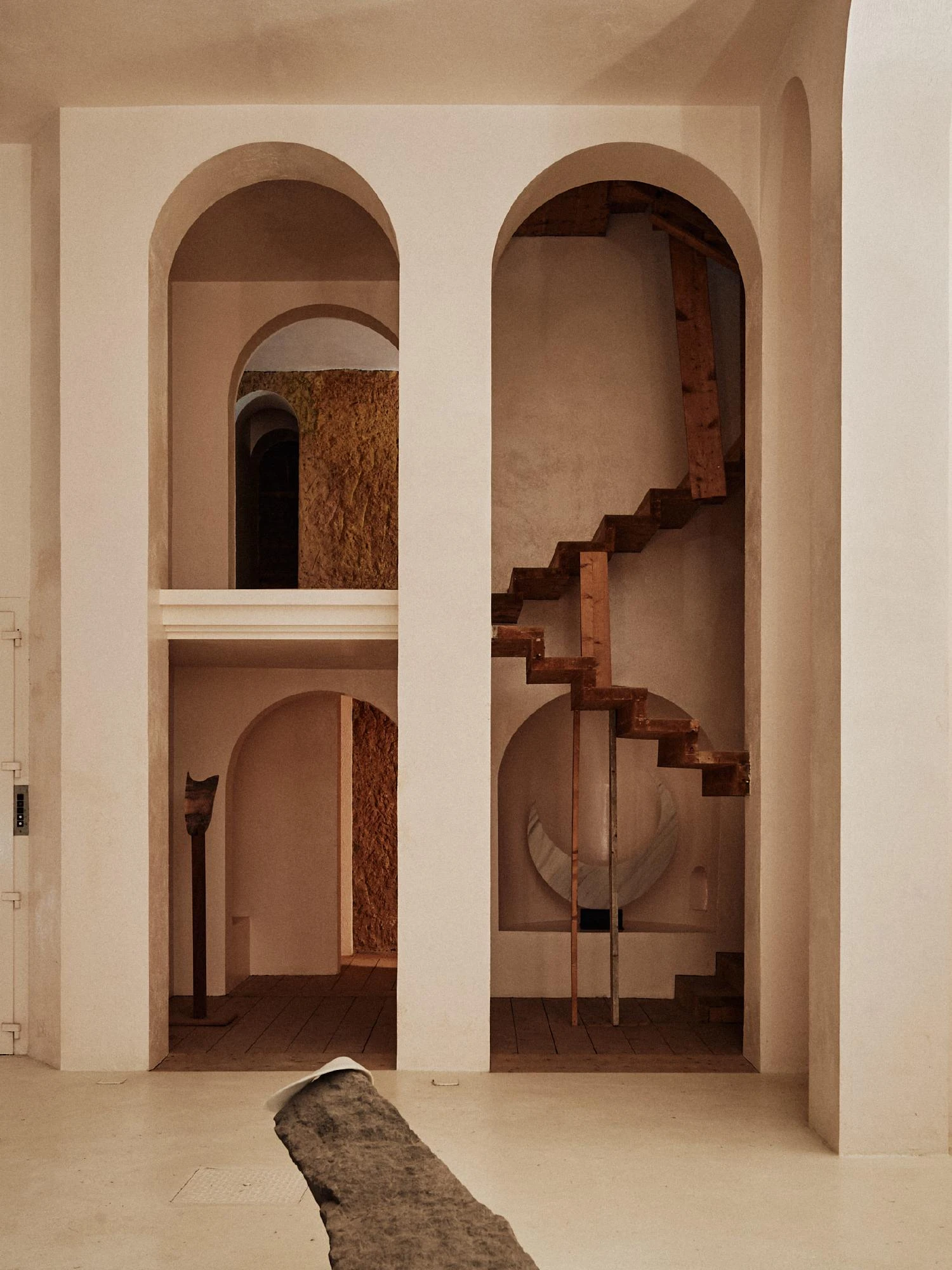 Xavier Corberó House and Studio by Xavier Corberó, photographed by Daniel Schäfer — Twin tall plaster arches frame a floating timber staircase and a suspended pale crescent form, a rolled stone slab running across the pale floor.