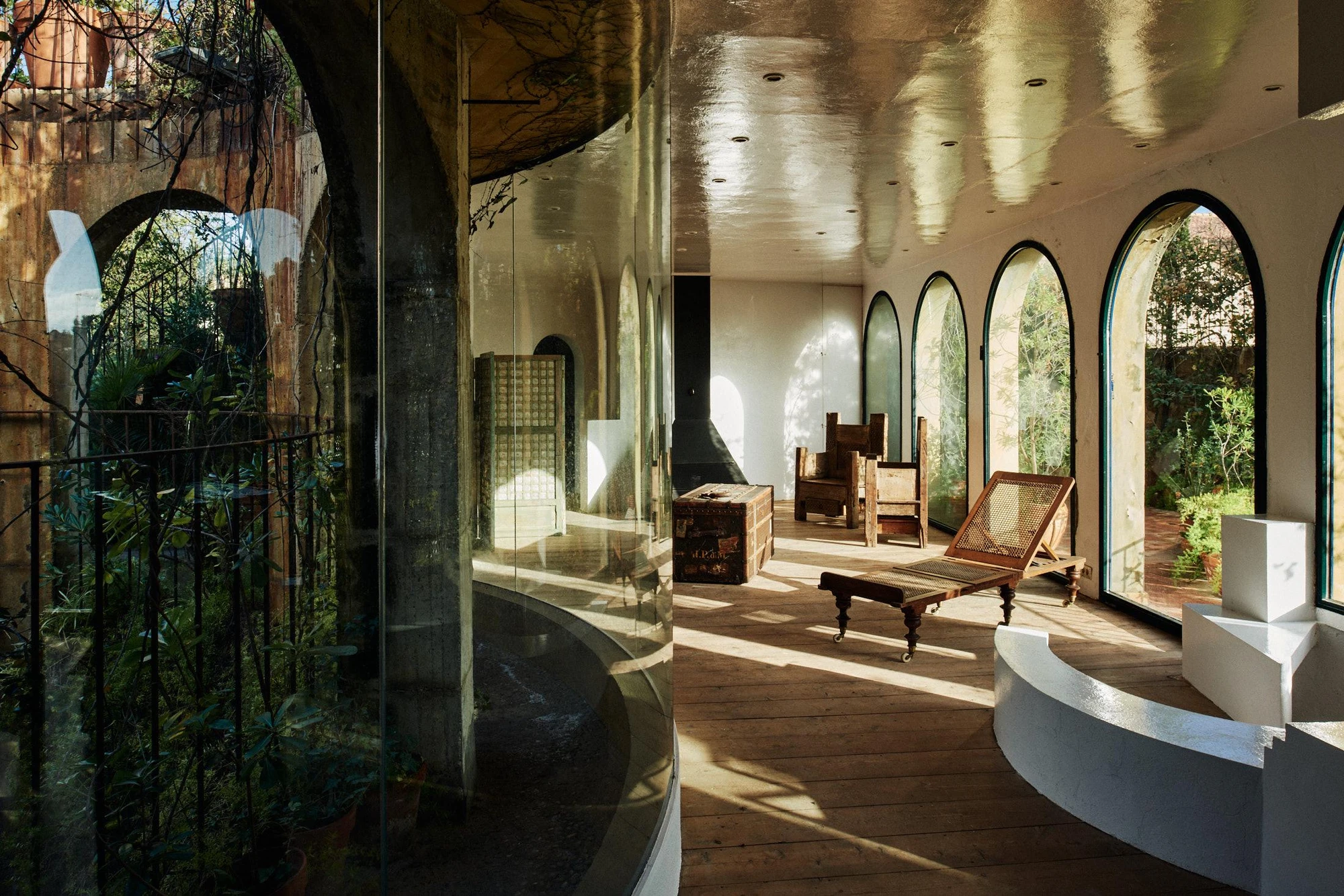 Xavier Corberó House and Studio by Xavier Corberó, photographed by Daniel Schäfer — A curved glazed gallery with pine plank floor and metallic ceiling holds a campaign chair and folding deck chair, the garden pressing at the arched glass.