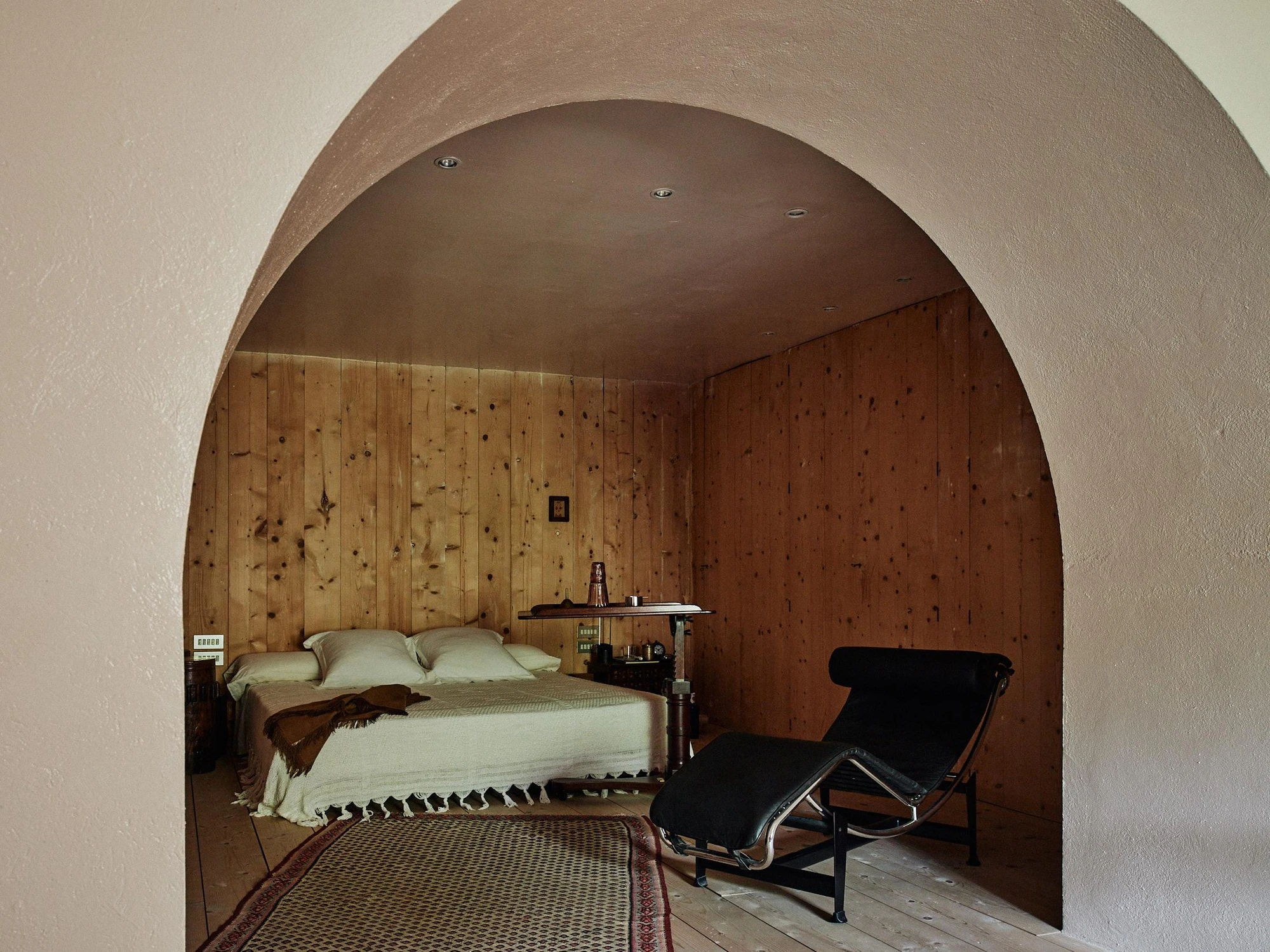 Xavier Corberó House and Studio by Xavier Corberó, photographed by Daniel Schäfer — A soft pink plaster arch frames a pine-lined bedroom where a low bed faces a black Le Corbusier LC4 chaise on a woven rug.