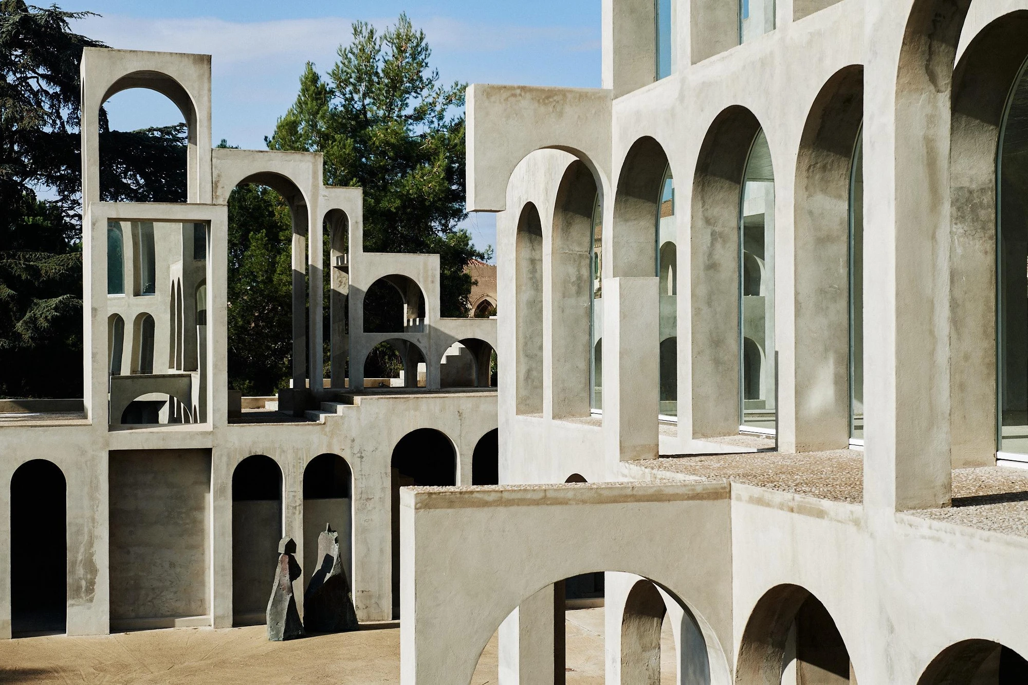 Xavier Corberó House and Studio by Xavier Corberó, photographed by Daniel Schäfer — Tiered concrete arcades step down into a gravel courtyard, a white chair and two small figures giving scale to the arched cliff of the compound.