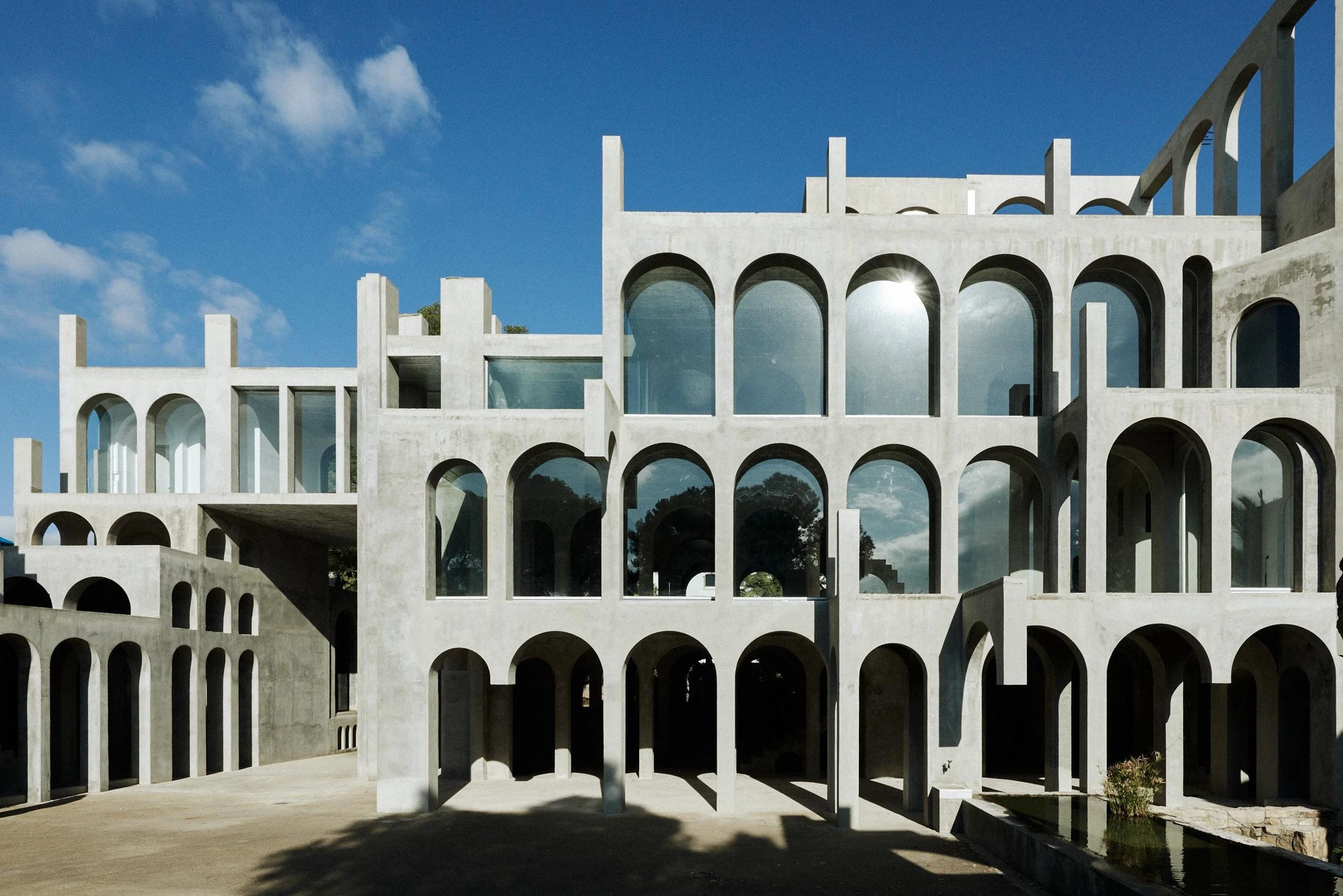 Xavier Corberó House and Studio by Xavier Corberó, photographed by Daniel Schäfer — A wide view of arcades wrapping a sunken courtyard and its long reflecting pool, umbrella pines rising beyond the concrete walls.