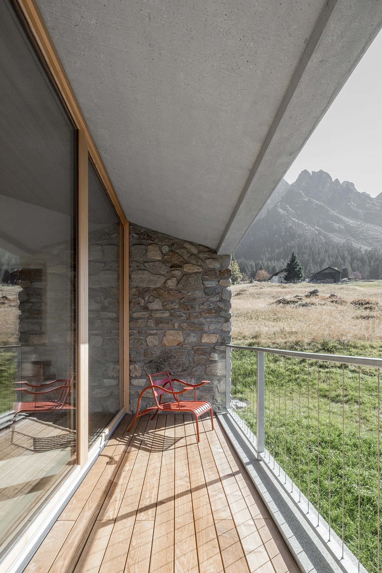 Vergalderie by Christian Tonko, photographed by Gustav Willeit — The balcony's larch deck under a board-formed concrete soffit, red steel chairs facing the peaks past a stone pier