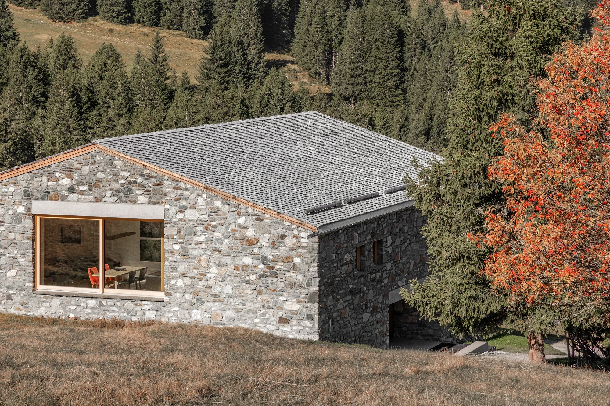 Vergalderie by Christian Tonko, photographed by Gustav Willeit — The full spruce-shingle roof seen from above, a wide window revealing red chairs inside and a darker stone service volume below
