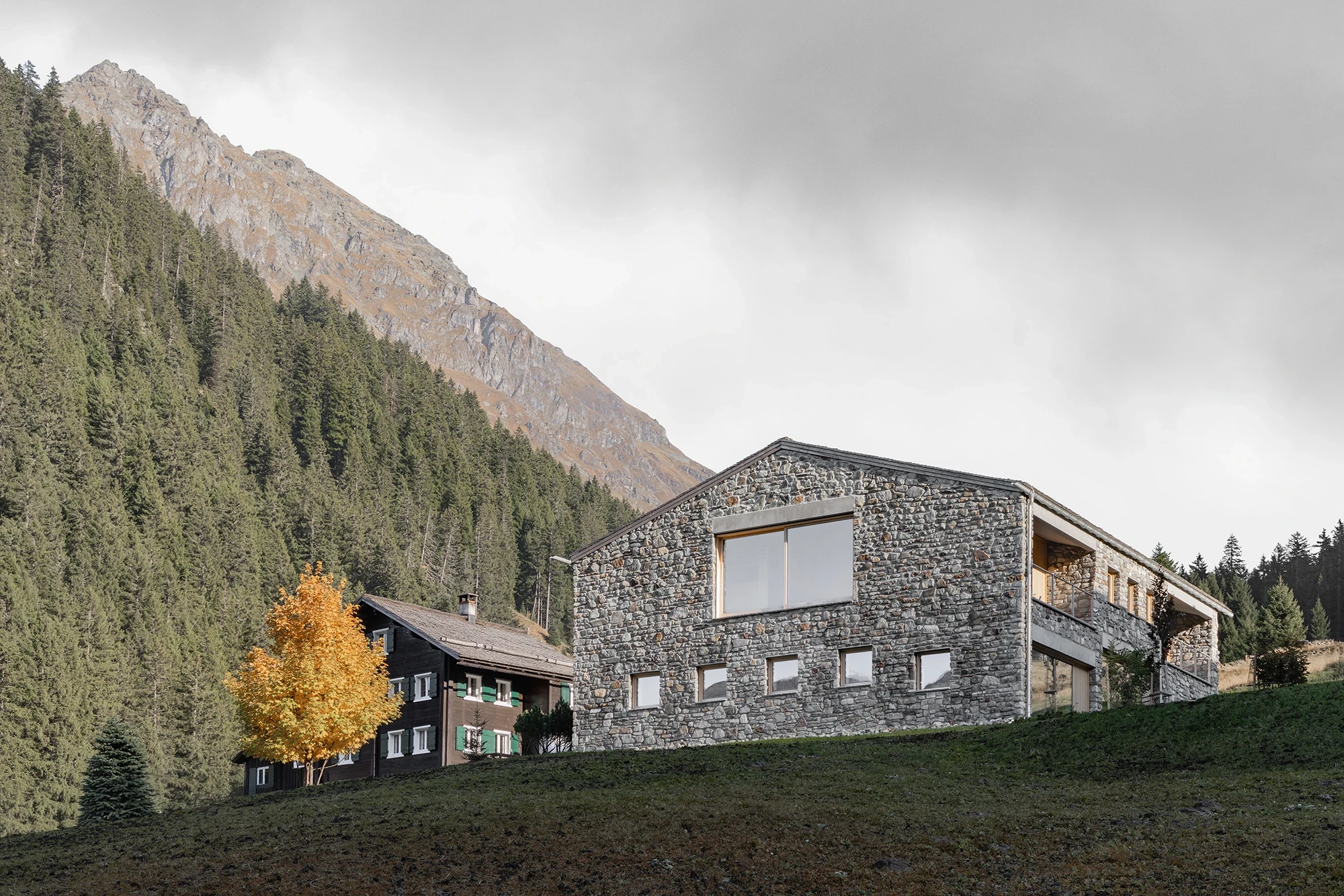 Vergalderie by Christian Tonko, photographed by Gustav Willeit — The stone volume steps down the slope beside an older painted timber chalet, its shingle roof low against the forest
