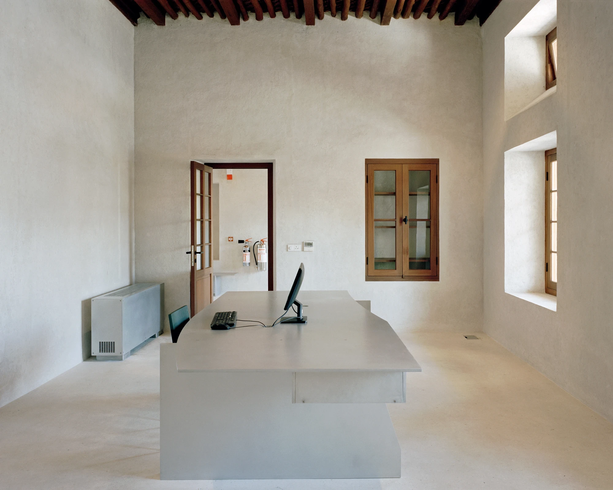 21 Customs House Post Office by Studio Anne Holtrop — Manama, Bahrain: a restored historic office with timber-beamed ceiling, wooden shutters and a pale concrete desk against white plaster walls.