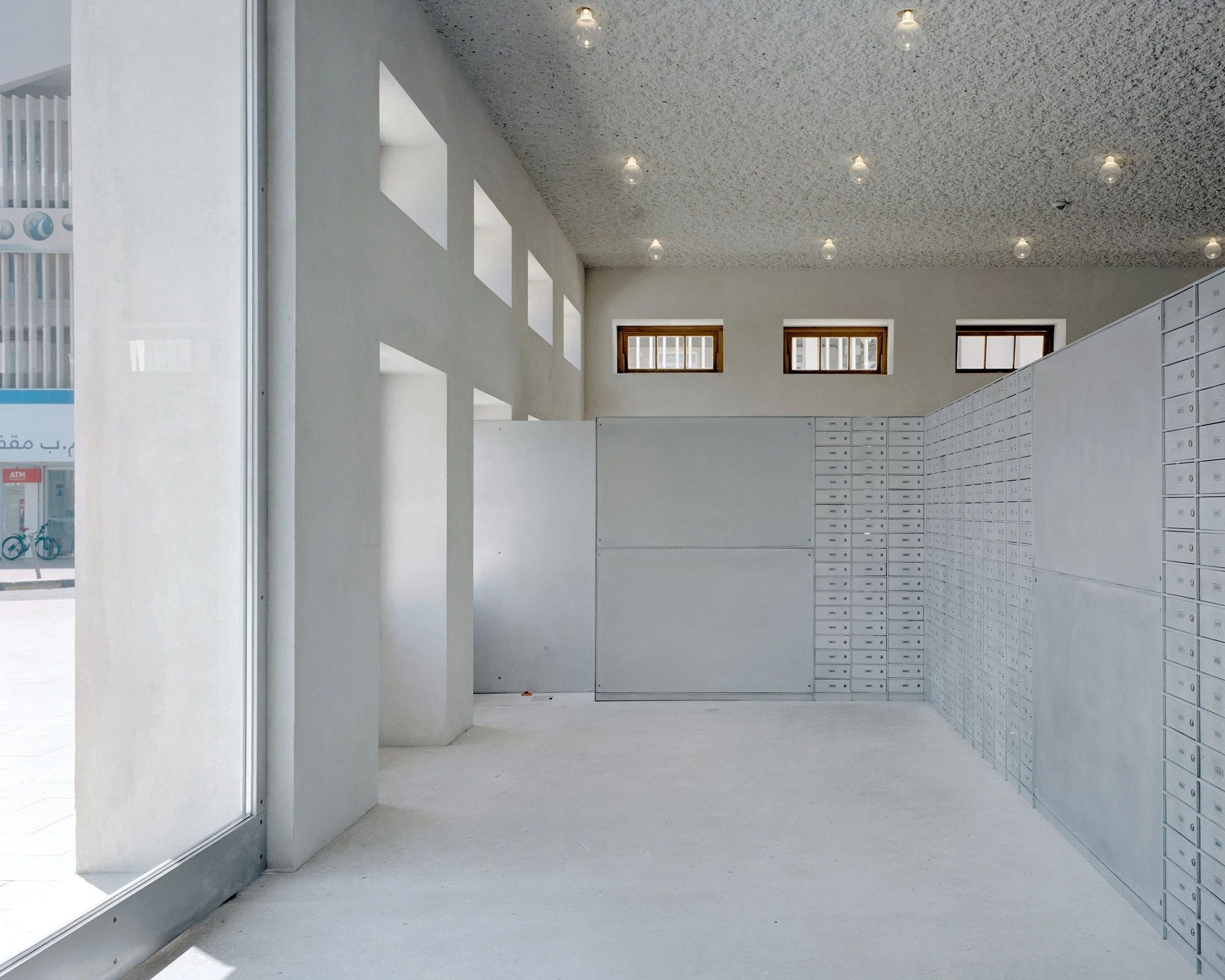 21 Customs House Post Office by Studio Anne Holtrop — Manama, Bahrain: a ground-floor room of grey post boxes lit through full-height glazing, small timber-shuttered windows set high in the concrete wall.