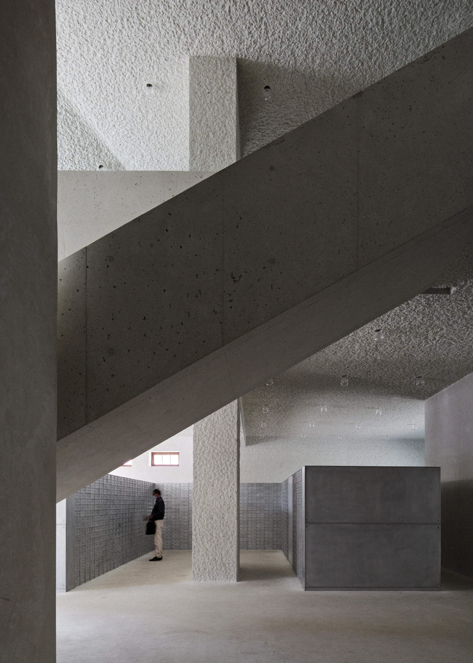 21 Customs House Post Office by Studio Anne Holtrop — Manama, Bahrain: a diagonal concrete beam crosses a double-height hall lined with post boxes, a lone visitor at the far wall for scale.