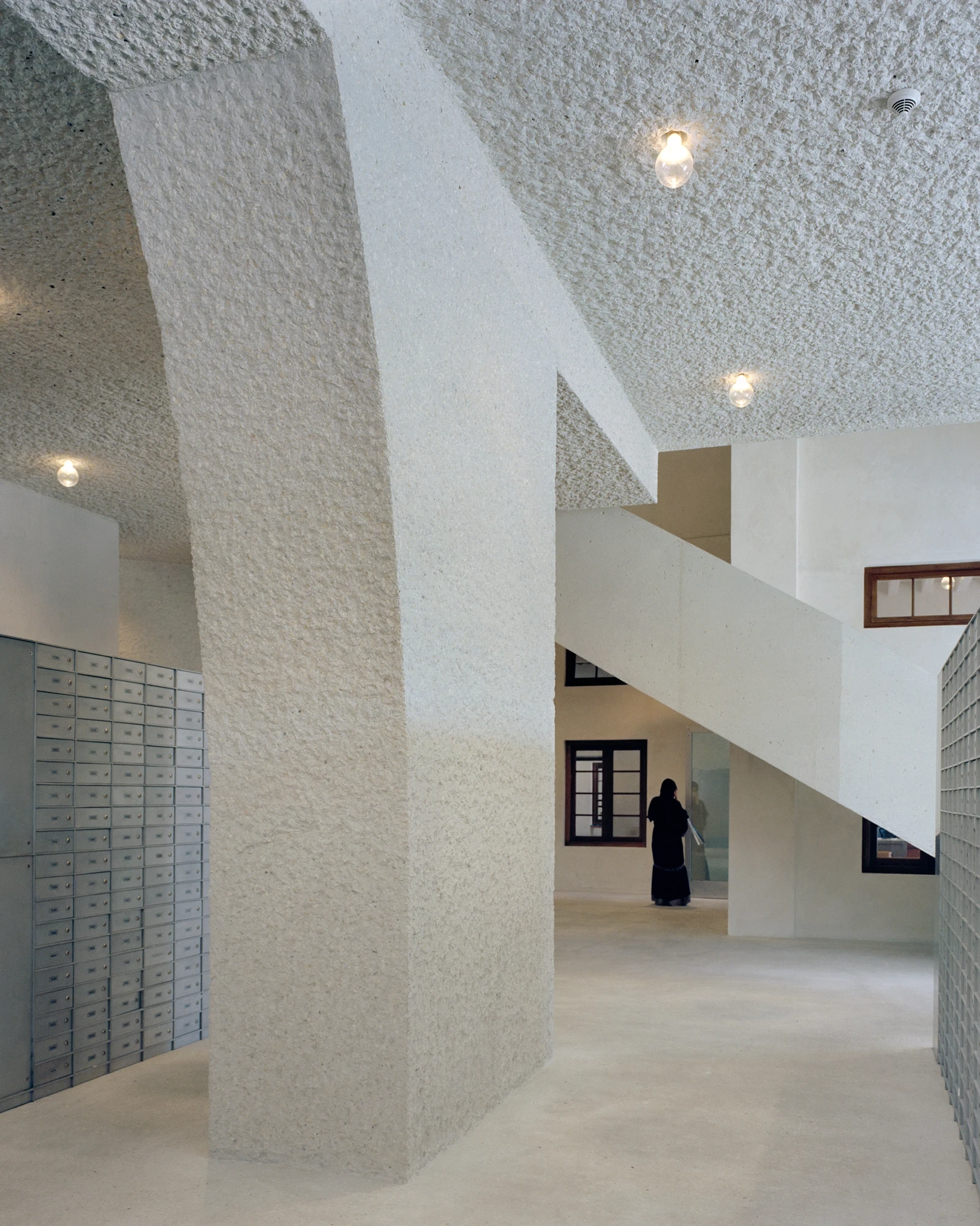 21 Customs House Post Office by Studio Anne Holtrop — Manama, Bahrain: a leaning pier of hand-hammered concrete meets a stair and a timber-framed door, a figure in black beyond.