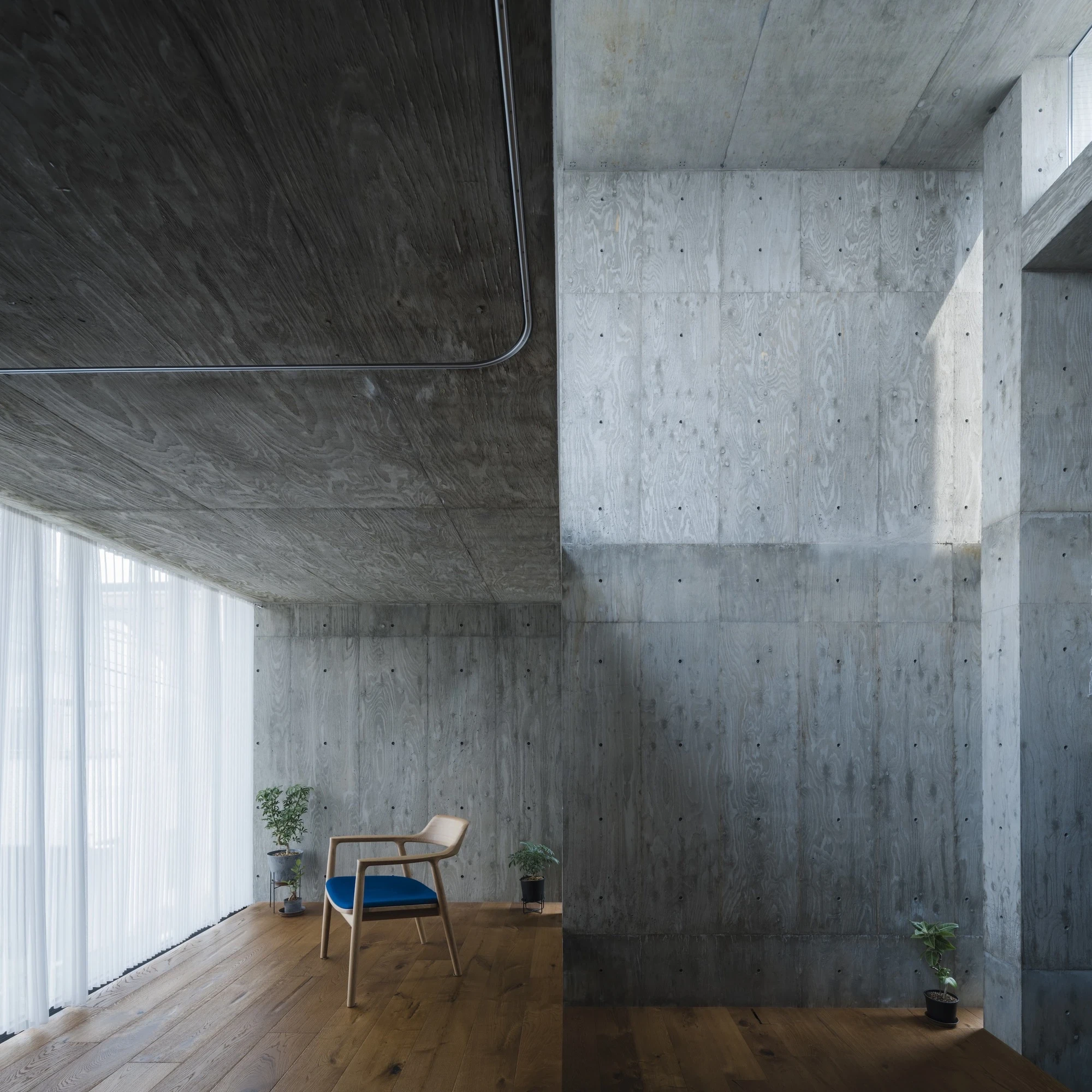 Building Frame of the House by IGArchitects, Japan — a bare concrete corner with a lone wood-and-fabric chair, oak floor and potted plants against board-formed walls.