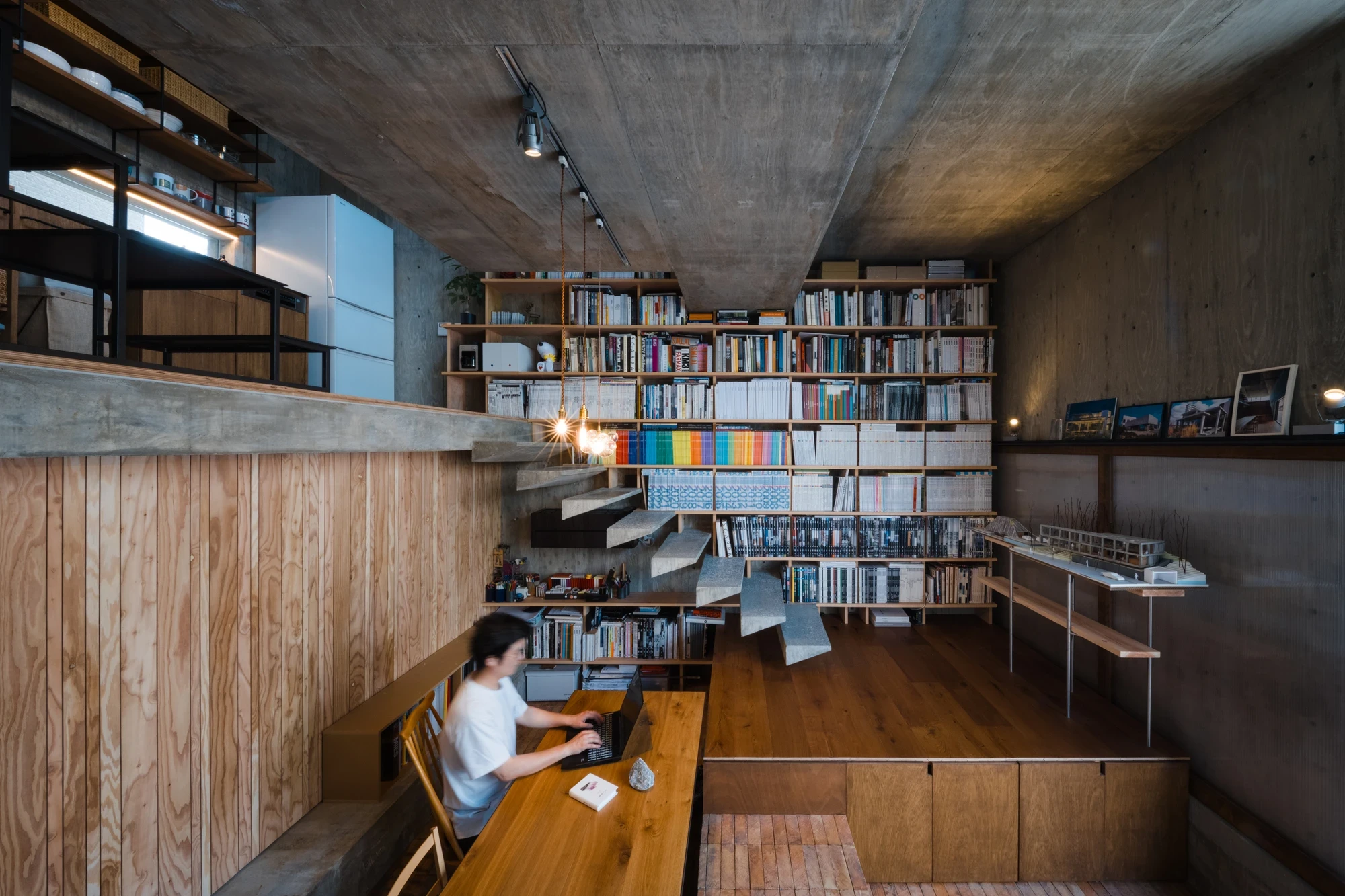 Building Frame of the House by IGArchitects, Japan — a resident works at a long oak desk below cantilevered concrete treads and a book wall, with offset slabs stacking overhead.