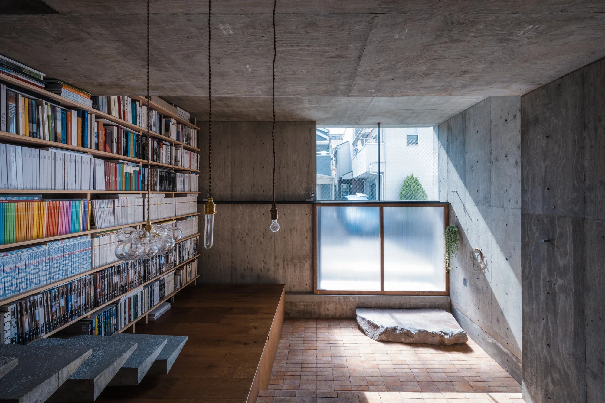 Building Frame of the House by IGArchitects, Japan — a full-height plywood-grained concrete room lined with a timber bookcase, a terracotta-brick floor and a stone slab set by a frosted glass window.
