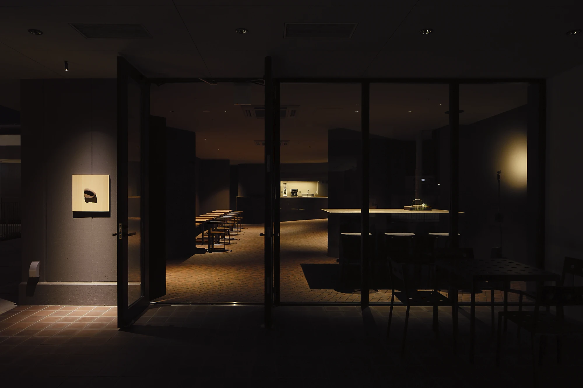 NIWA FUKUOKA by ANONIMAL design community — Fukuoka, Japan: seen at night through the glass facade, the warm-lit interior glows against dark tile, a small framed work lit beside the entrance door.