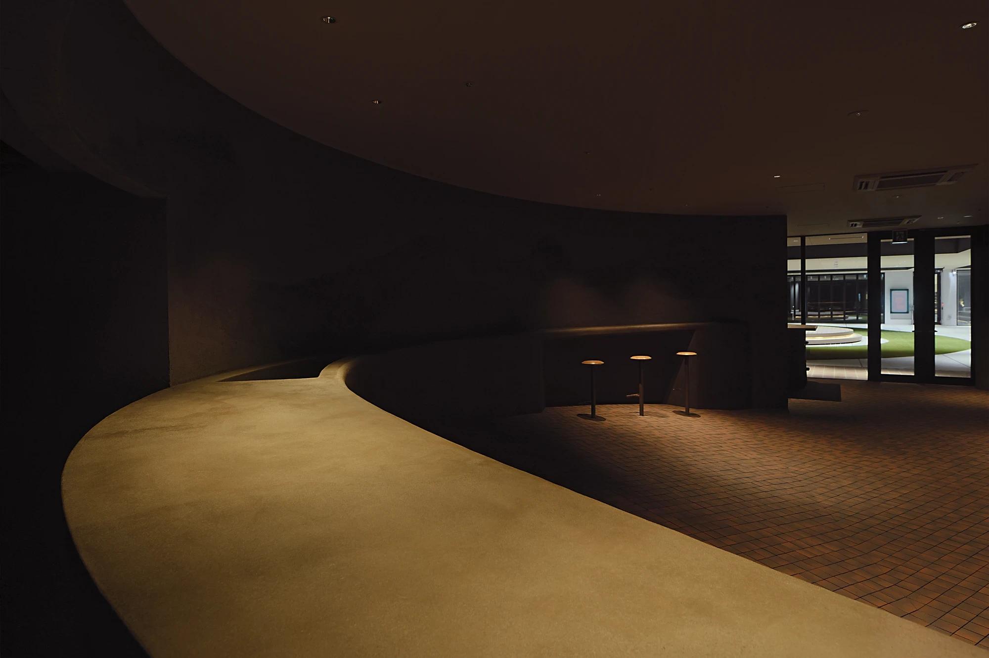 NIWA FUKUOKA by ANONIMAL design community — Fukuoka, Japan: the raised earthen stage sweeps in a long curve above the terracotta floor, its warm surface caught in a single band of light.