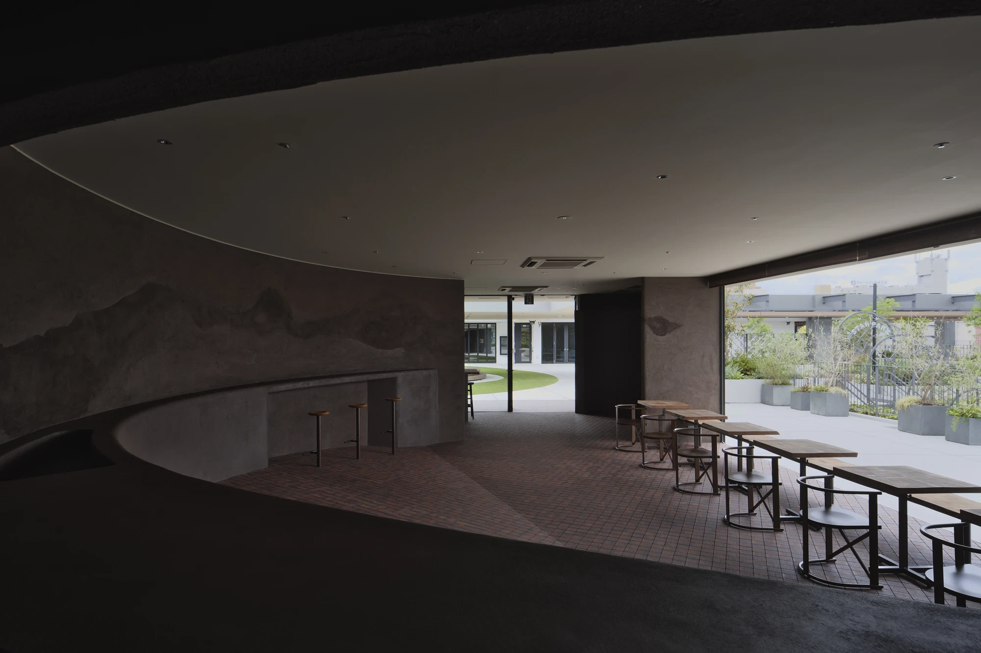 NIWA FUKUOKA by ANONIMAL design community — Fukuoka, Japan: the curved earthen room opens through full-height glazing onto the daylit rooftop plaza, terracotta tile floor meeting the pale paving beyond.
