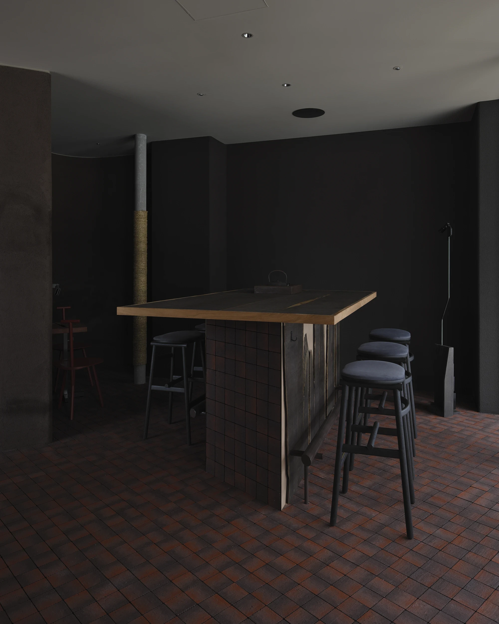 NIWA FUKUOKA by ANONIMAL design community — Fukuoka, Japan: a timber-topped bar counter on a tiled base stands against a near-black wall, flanked by upholstered stools.