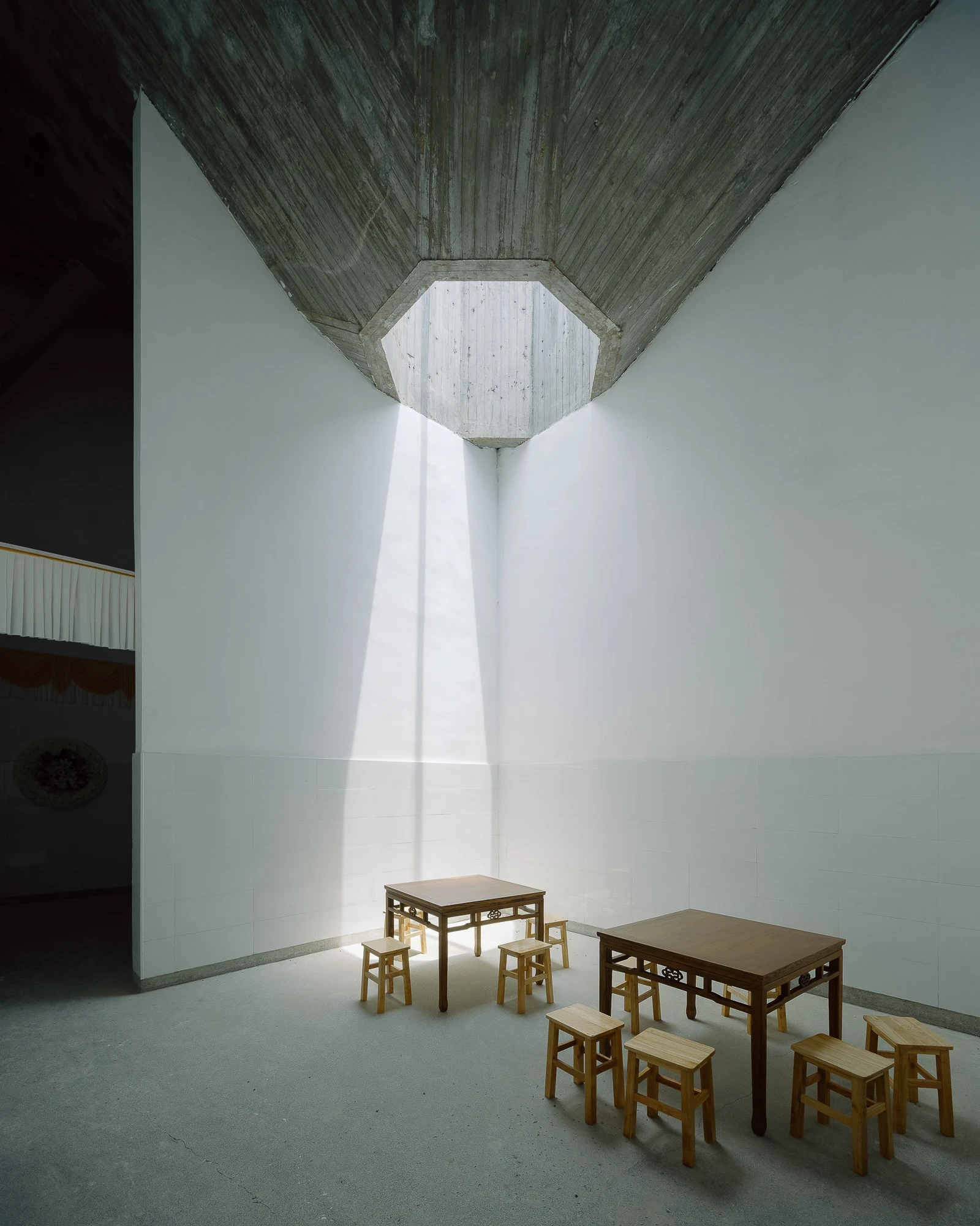Suichang Funerary Complex by Untitled Architects, photographed by Tianzhou Yang — a shaft of daylight falls through an octagonal opening onto plain timber tables and stools in a white-walled room.