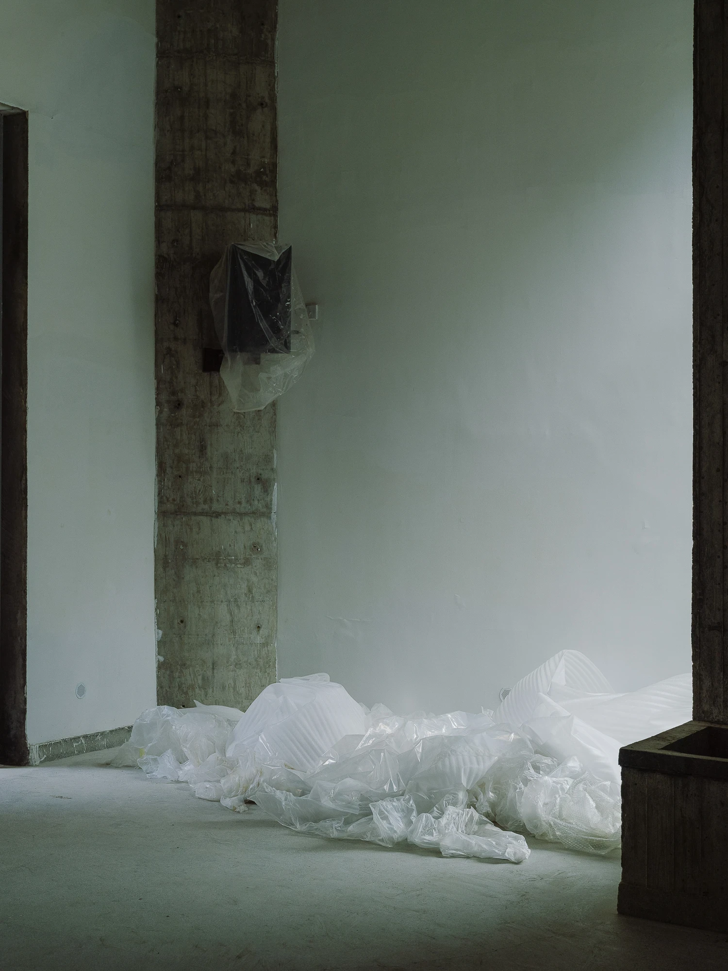 Suichang Funerary Complex by Untitled Architects, photographed by Tianzhou Yang — a bare concrete room where plastic sheeting gathers on the floor beside a raw column and a dark timber crate.