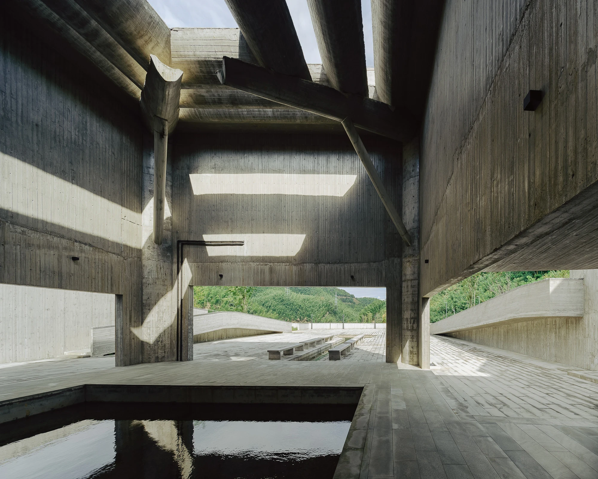 Suichang Funerary Complex by Untitled Architects, photographed by Tianzhou Yang — angled shafts of sunlight cross a concrete court open to the valley, a reflecting pool in the foreground.
