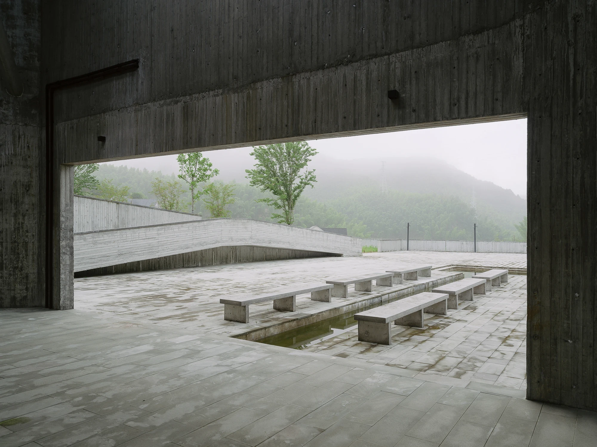 Suichang Funerary Complex by Untitled Architects, photographed by Tianzhou Yang — a covered edge looks out over a terrace of concrete benches straddling a water channel, misted hills beyond.