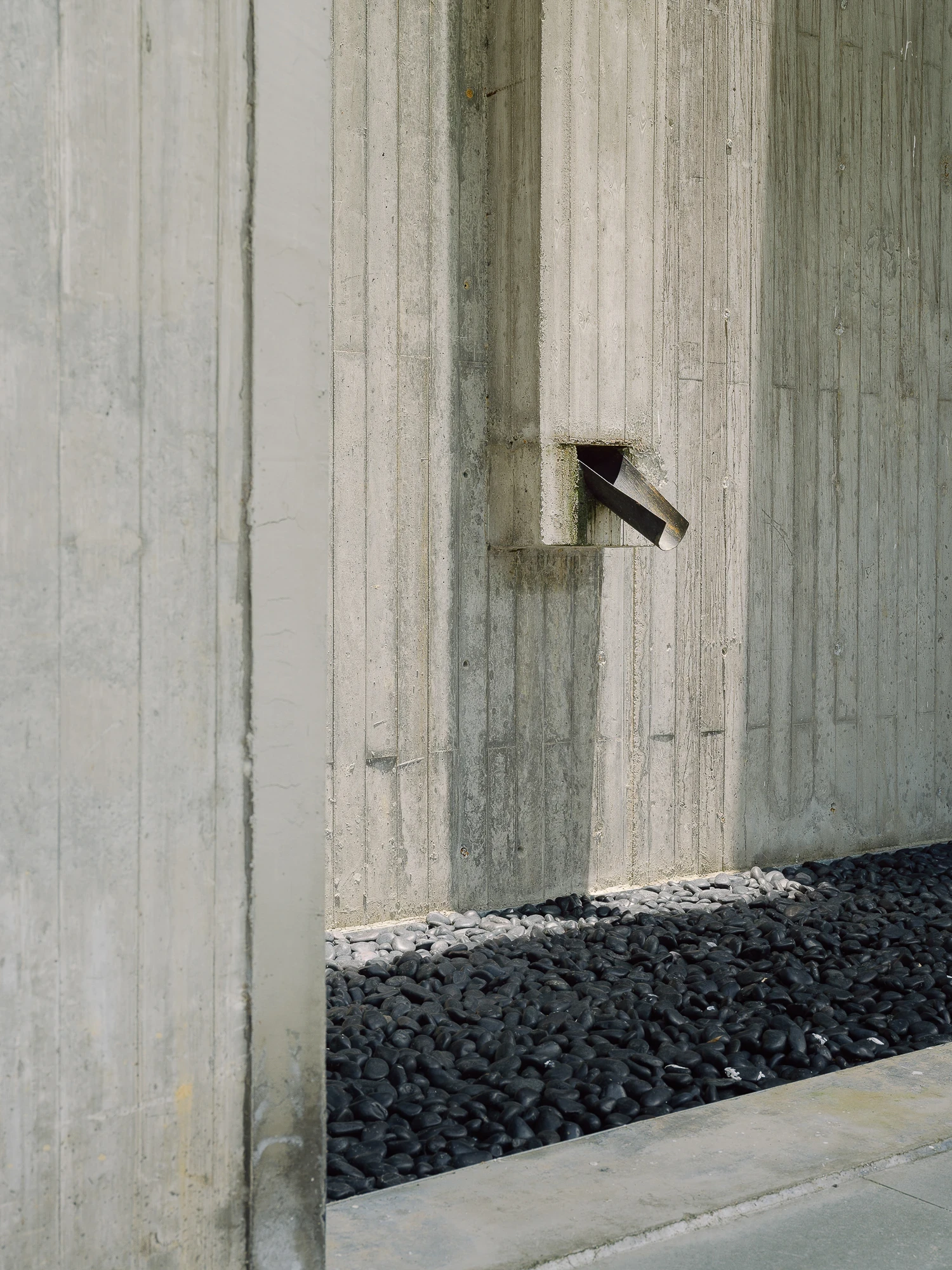 Suichang Funerary Complex by Untitled Architects, photographed by Tianzhou Yang — a steel spout set into a board-formed concrete wall drips onto a bed of black river stones.
