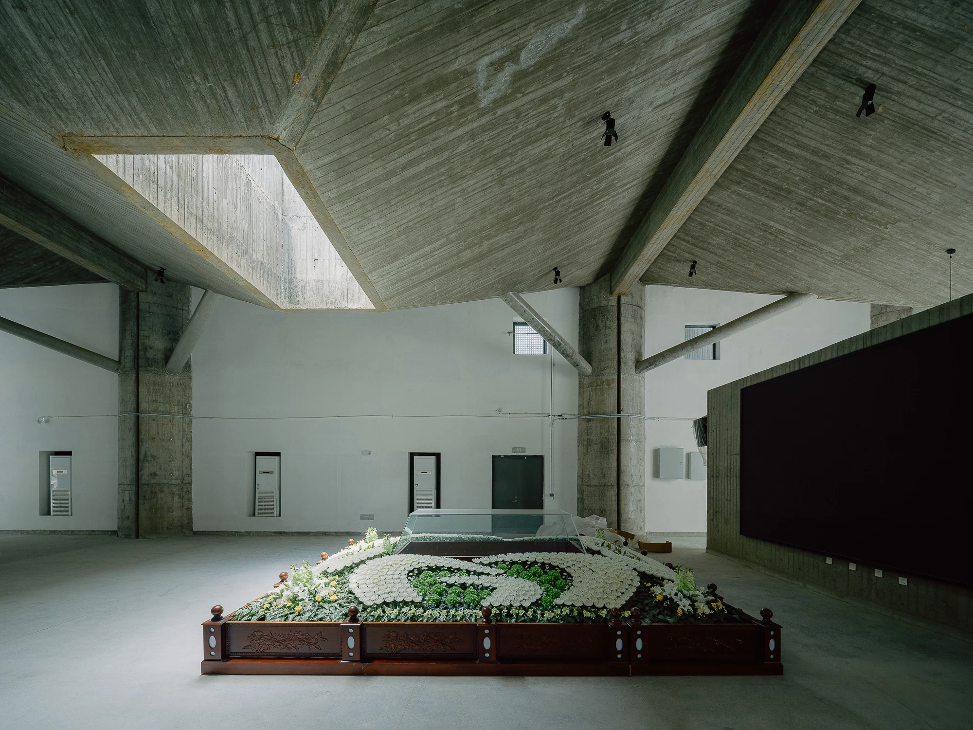 Suichang Funerary Complex by Untitled Architects, photographed by Tianzhou Yang — a coffin covered in white and green flowers beneath faceted concrete beams and a dark screen wall.