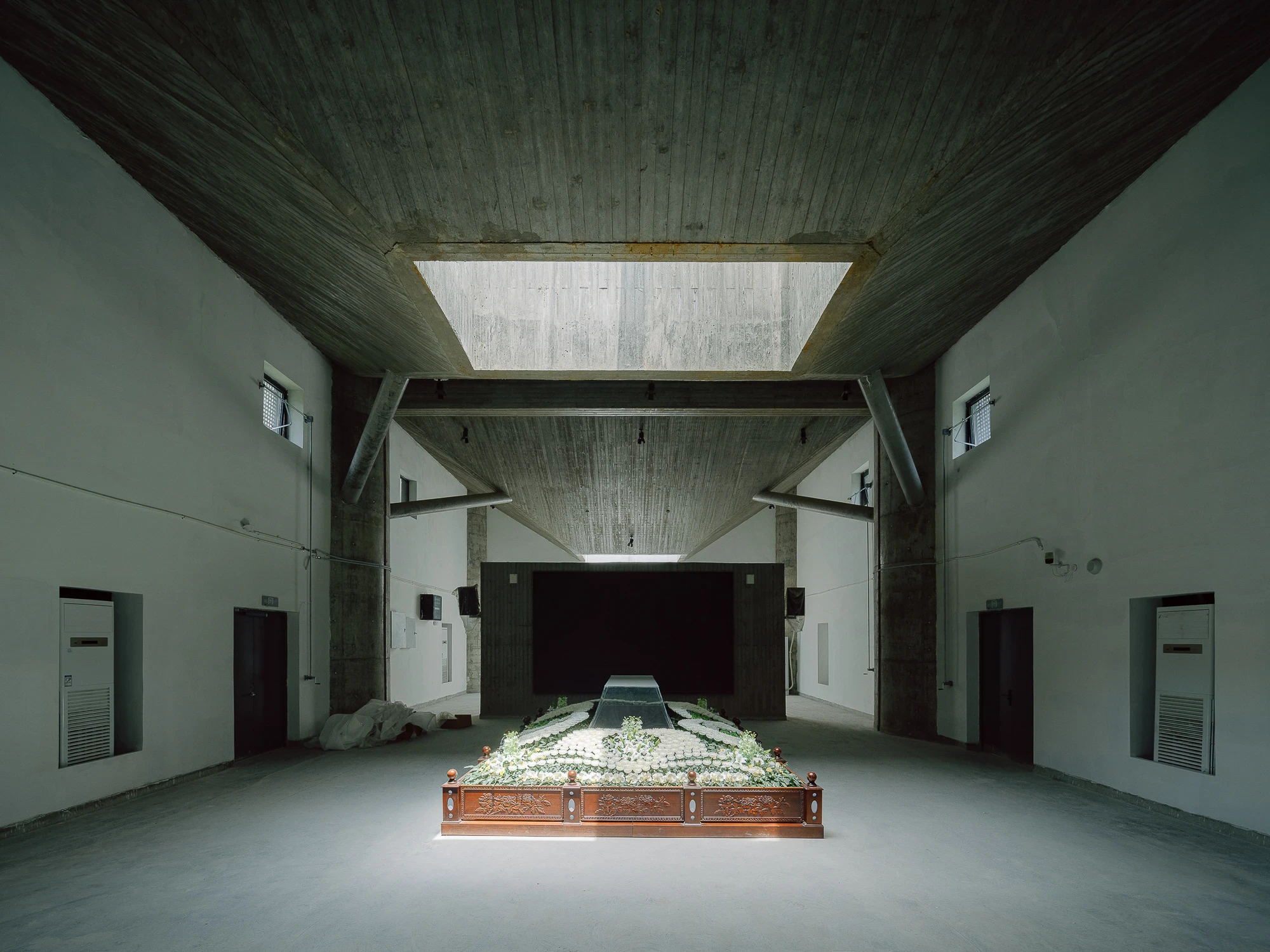 Suichang Funerary Complex by Untitled Architects, photographed by Tianzhou Yang — a tall mourning hall lit by a rectangular skylight, a flower-banked bier on a carved timber base at its centre.
