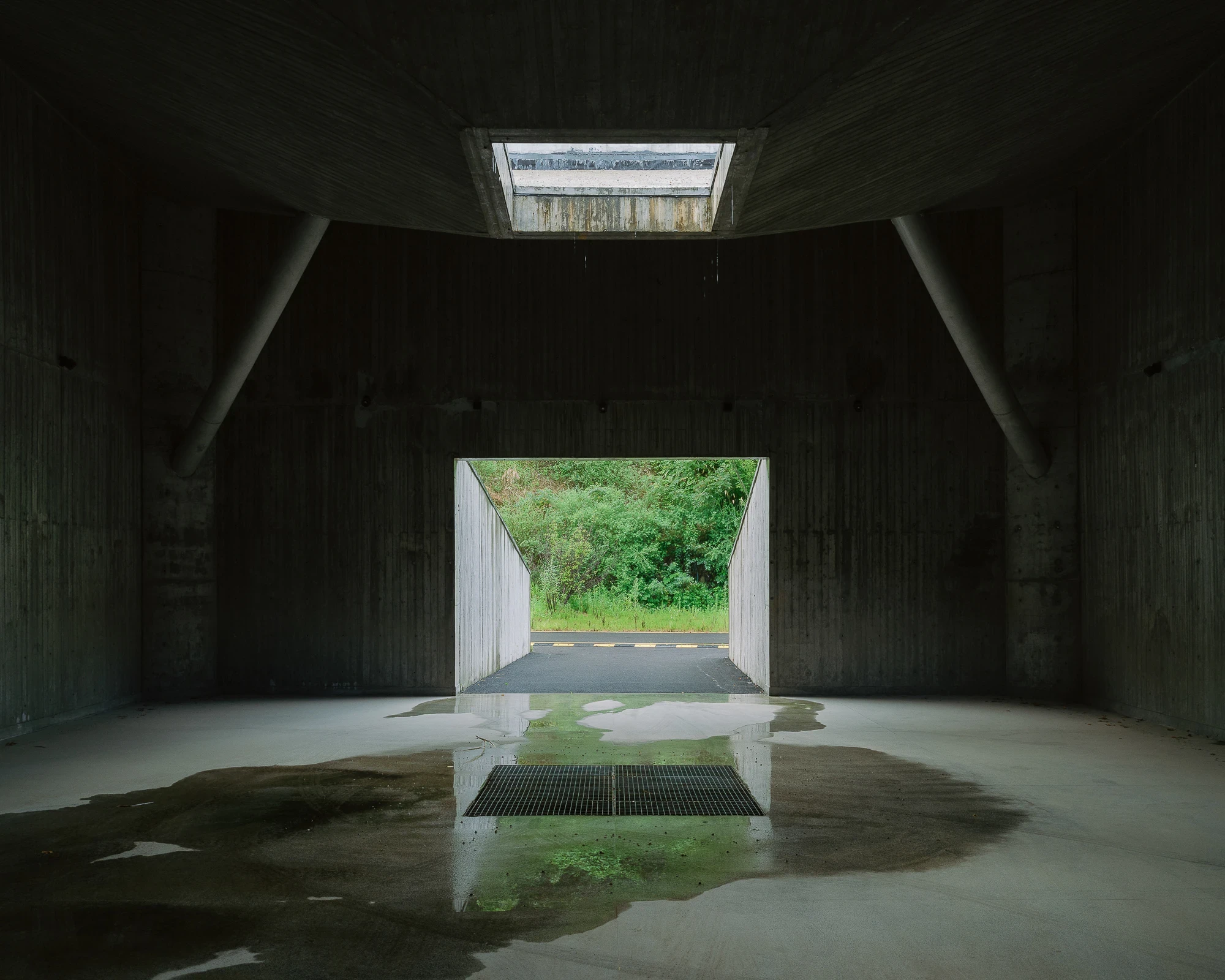 Suichang Funerary Complex by Untitled Architects, photographed by Tianzhou Yang — a dark concrete chamber opening onto a green hillside, a skylight and floor grate above pooled water.