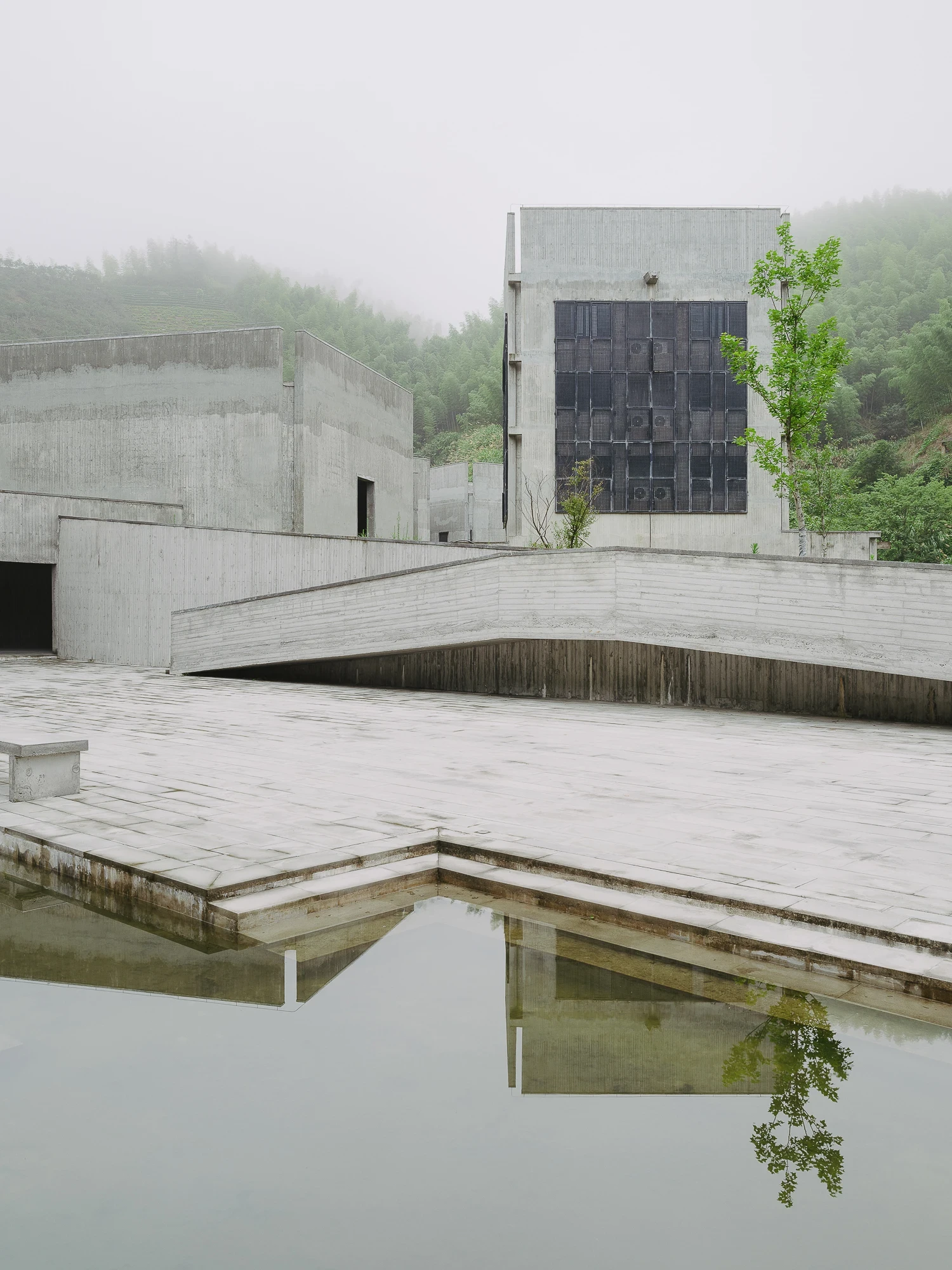 Suichang Funerary Complex by Untitled Architects, photographed by Tianzhou Yang — grey concrete volumes and a folded ramp step down to a reflecting pool under misted hills, a gridded window in the tallest block.