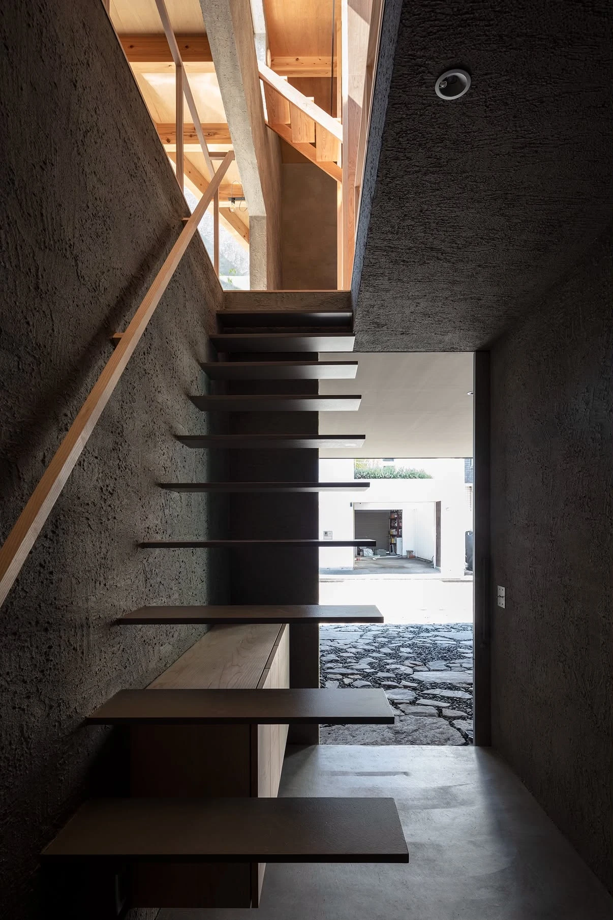 NesT by mA-style architects, photographed by Norihito Yamauchi — Dark steel stair treads float against near-black plaster, the open street doorway framing a stone-paved forecourt beyond.