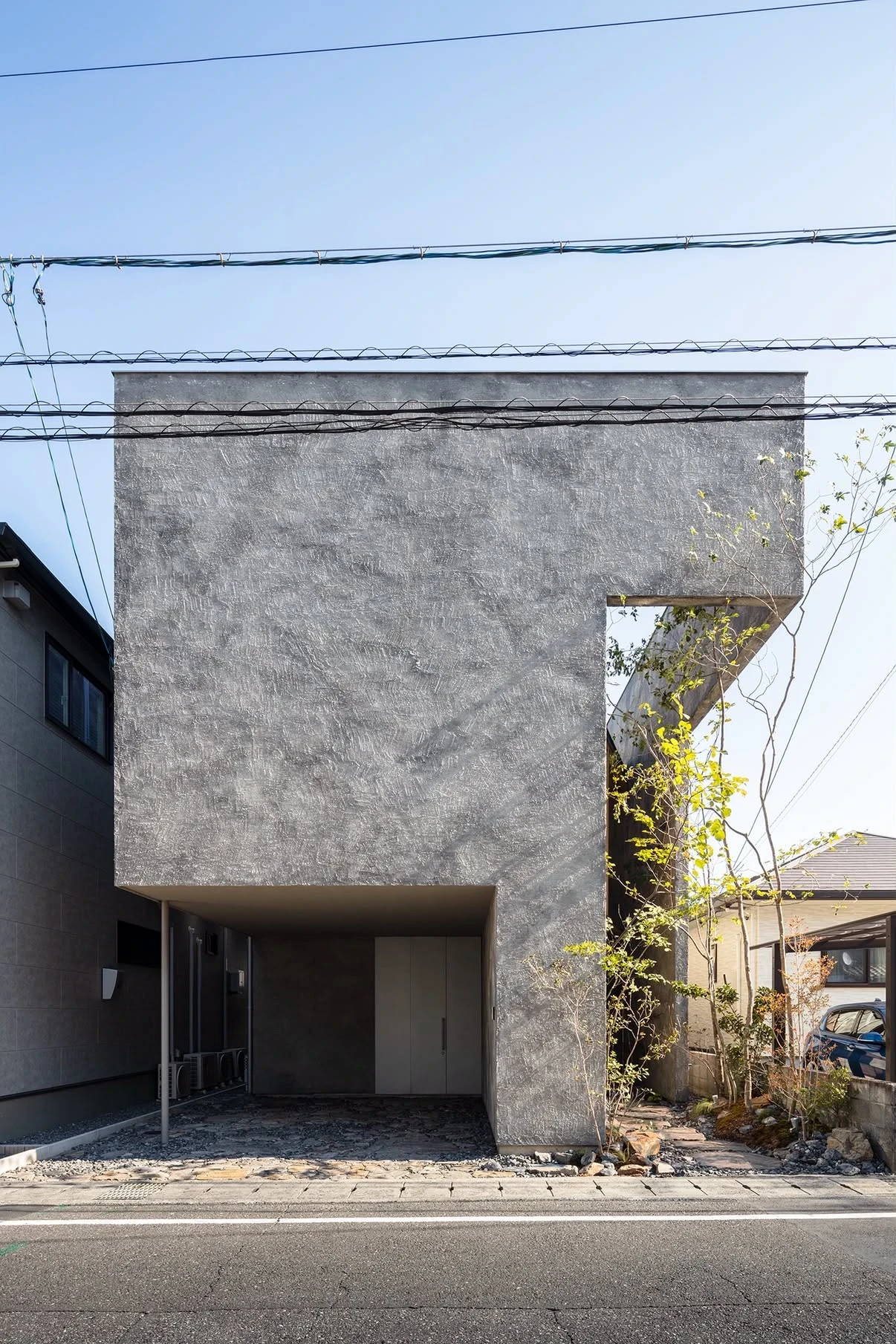 NesT by mA-style architects, photographed by Norihito Yamauchi — The closed grey facade seen head-on from the road, its flat mortar plane broken only by the carport recess and the tall planted slot.