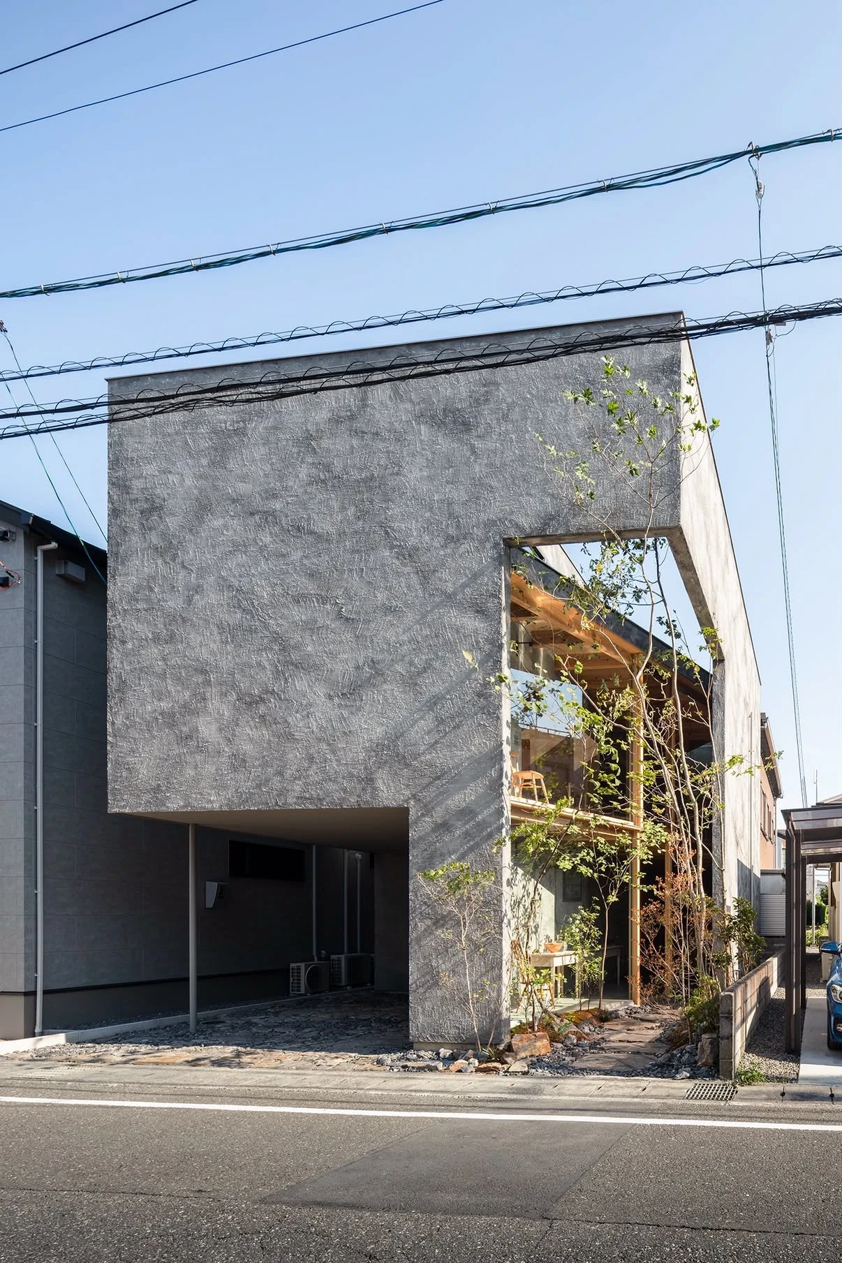 NesT by mA-style architects, photographed by Norihito Yamauchi — Morning light rakes the street facade, its grey mortar mass opened by a deep vertical cut where timber structure and a tree show through.
