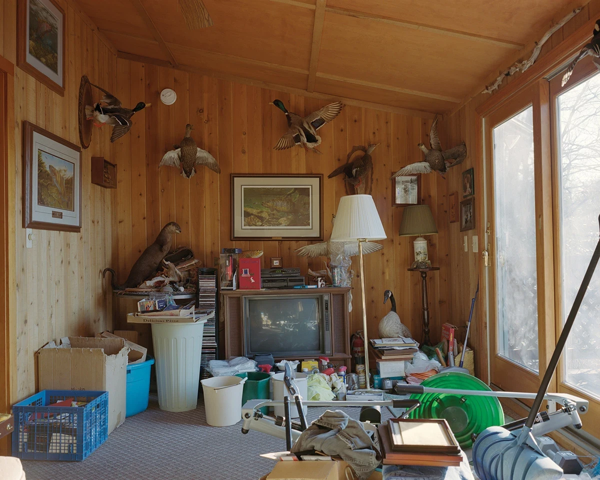 Wading by Jon Testa, photographed by Jon Testa — a cluttered pine-paneled sunroom hung with mounted ducks and a game fish, a boxy old television and exercise equipment crowding the floor.
