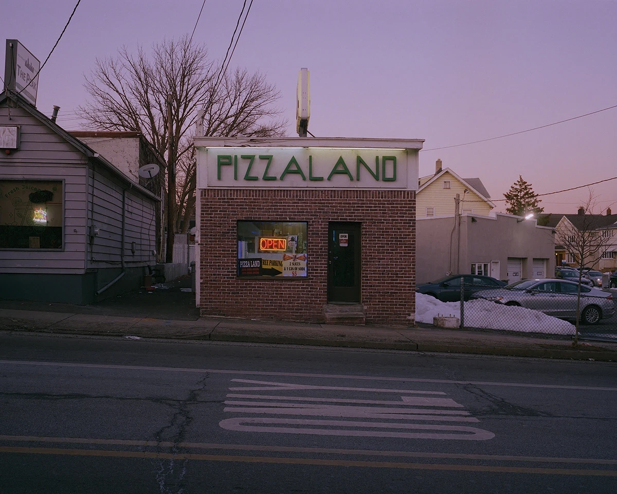 Wading by Jon Testa, photographed by Jon Testa — a brick Pizzaland storefront glowing green against a purple dusk sky, snowmelt and parked cars beside it.