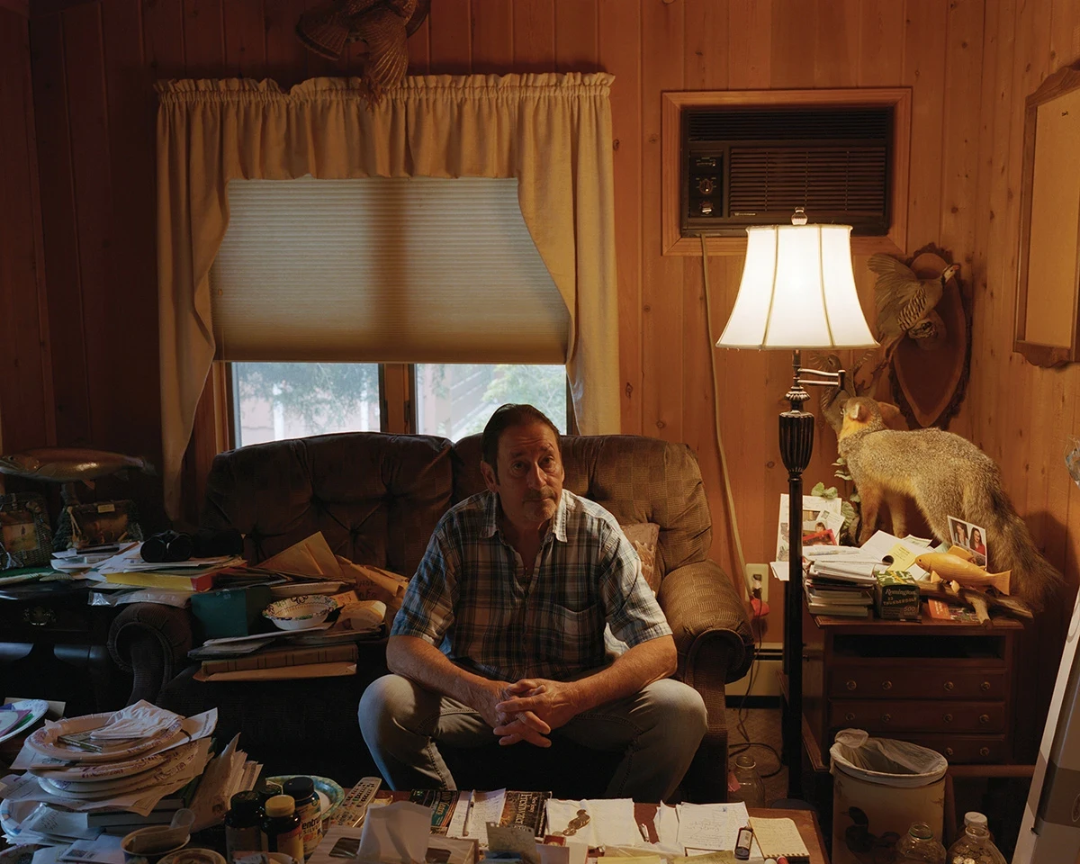 Wading by Jon Testa, photographed by Jon Testa — a man in a plaid shirt sits in a brown recliner ringed by taxidermy, a lit lamp, and tables buried in mail and pill bottles.