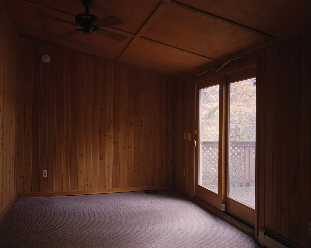 Wading by Jon Testa, photographed by Jon Testa — an empty pine-paneled room with dented pale carpet and a ceiling fan, a sliding glass door open onto yellowing autumn trees.