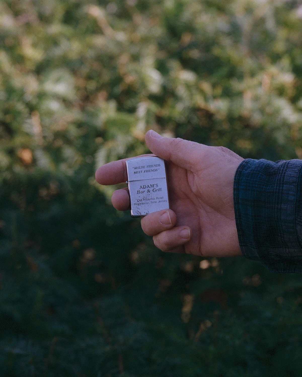 Wading by Jon Testa, photographed by Jon Testa — a hand in a plaid sleeve holds a silver Zippo lighter engraved for Adam's Bar & Grill against a blurred green hedge.