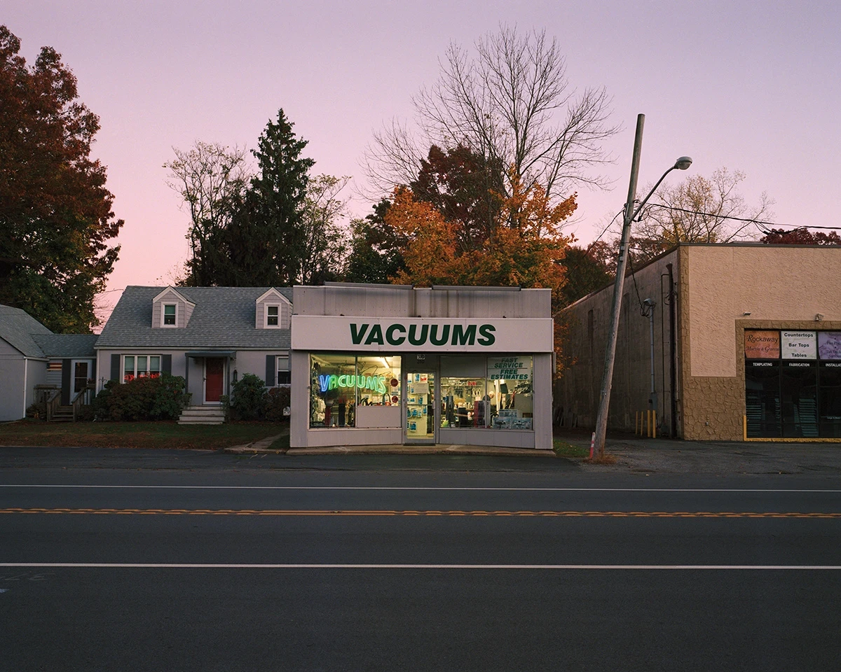 Wading by Jon Testa, photographed by Jon Testa — a small storefront reading VACUUMS beside a suburban house under a pink and lavender dusk sky.