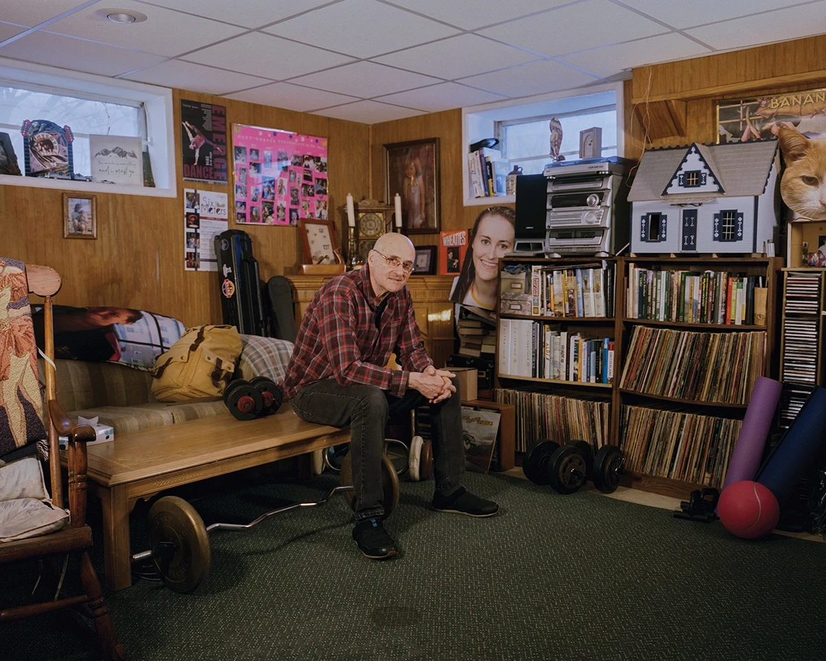 Wading by Jon Testa, photographed by Jon Testa — a man and a younger woman in a wood-paneled basement lined with records, books, and dumbbells, a dollhouse on a shelf behind them.