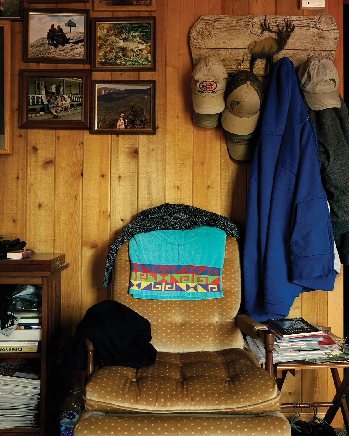 Wading by Jon Testa, photographed by Jon Testa — a worn upholstered chair draped with a patterned towel and jacket below a pine wall of framed family photographs and hanging ball caps.