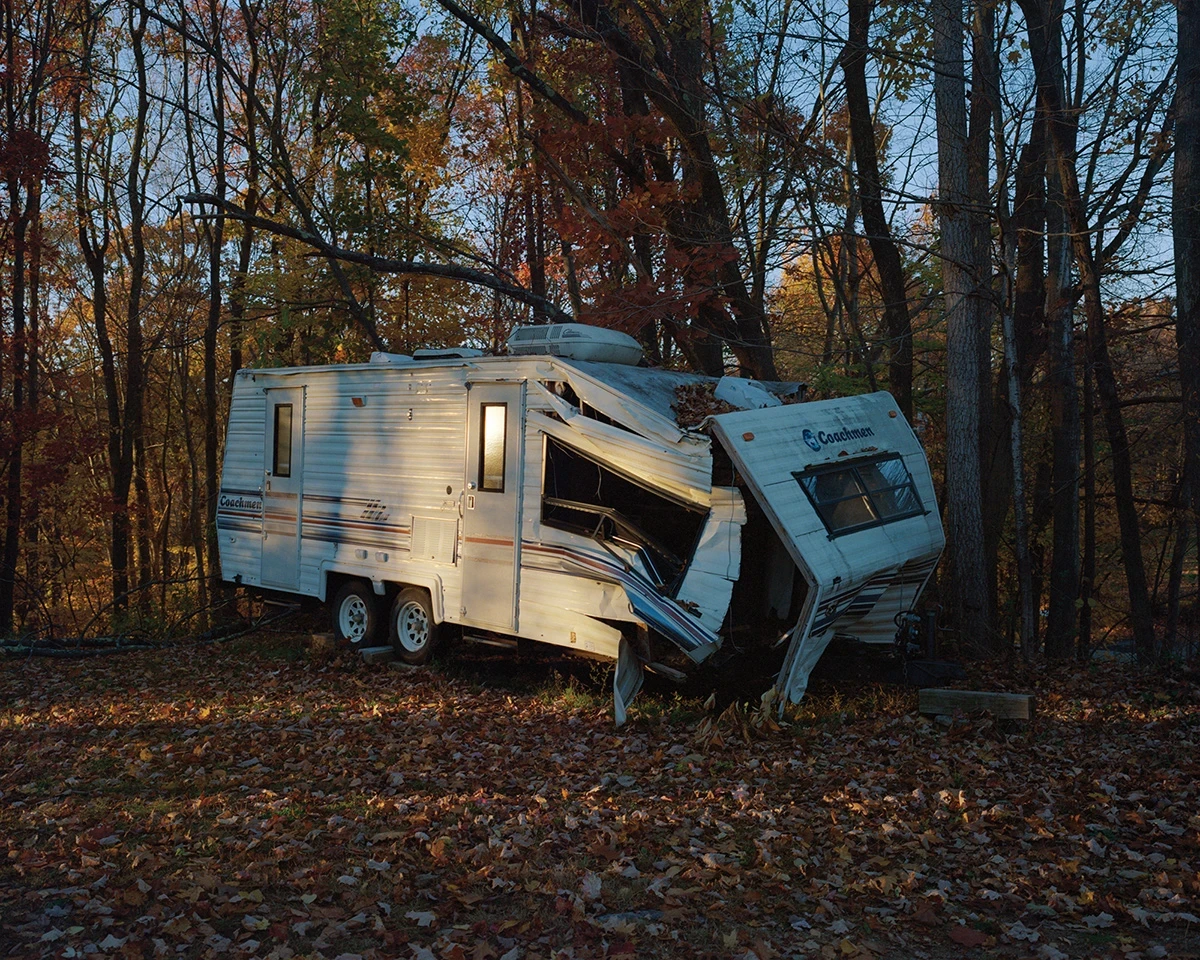 Wading by Jon Testa, photographed by Jon Testa — a white Coachman travel camper split open and rusting at the edge of a leaf-covered clearing in bare autumn woods at dusk.