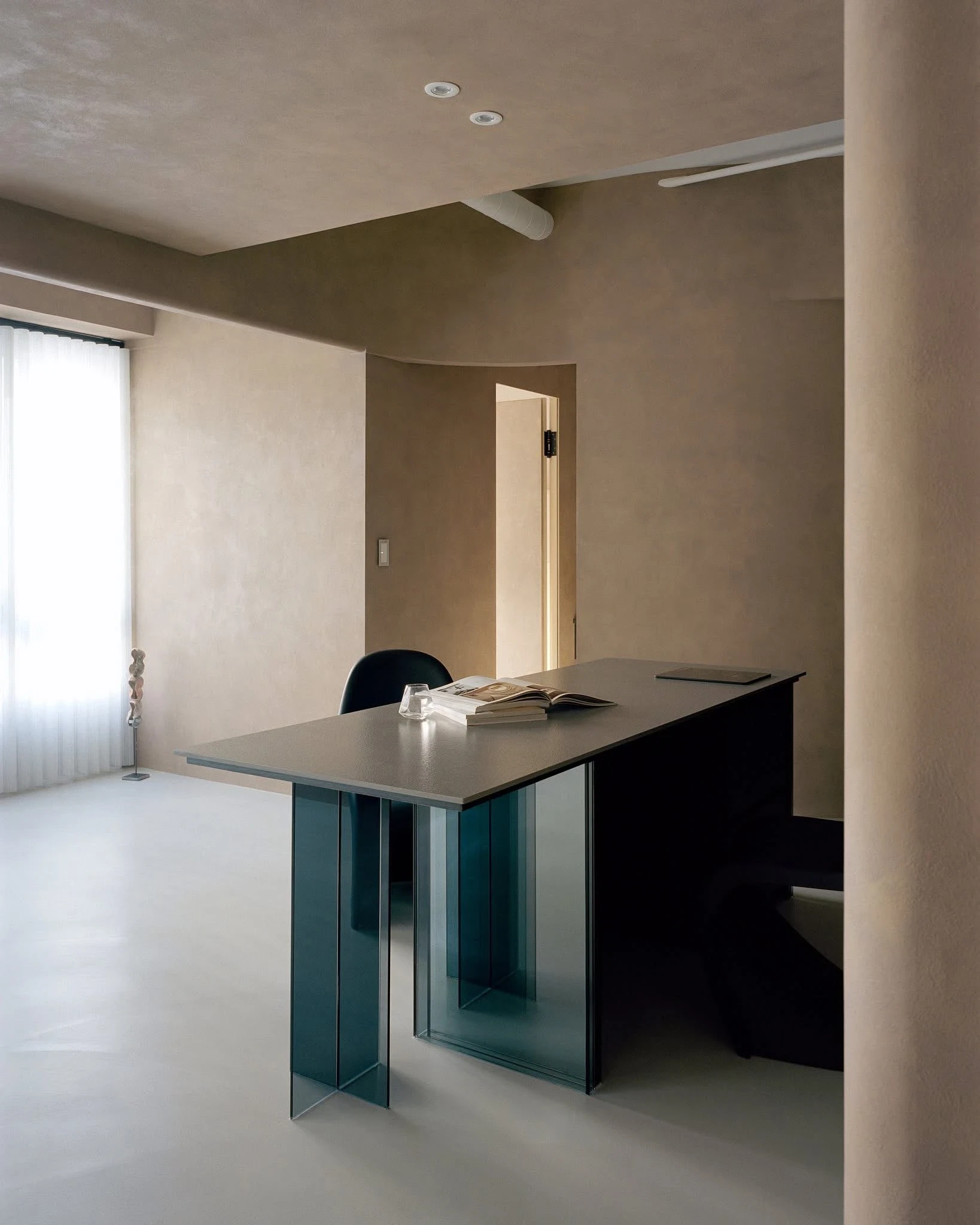 Uno's Purr-fect Time by W&Li Design, photographed by Studio Millspace — the blue-grey glass dining table seen against a rounded plaster wall and a doorway, its transparent legs catching the grey-green floor.