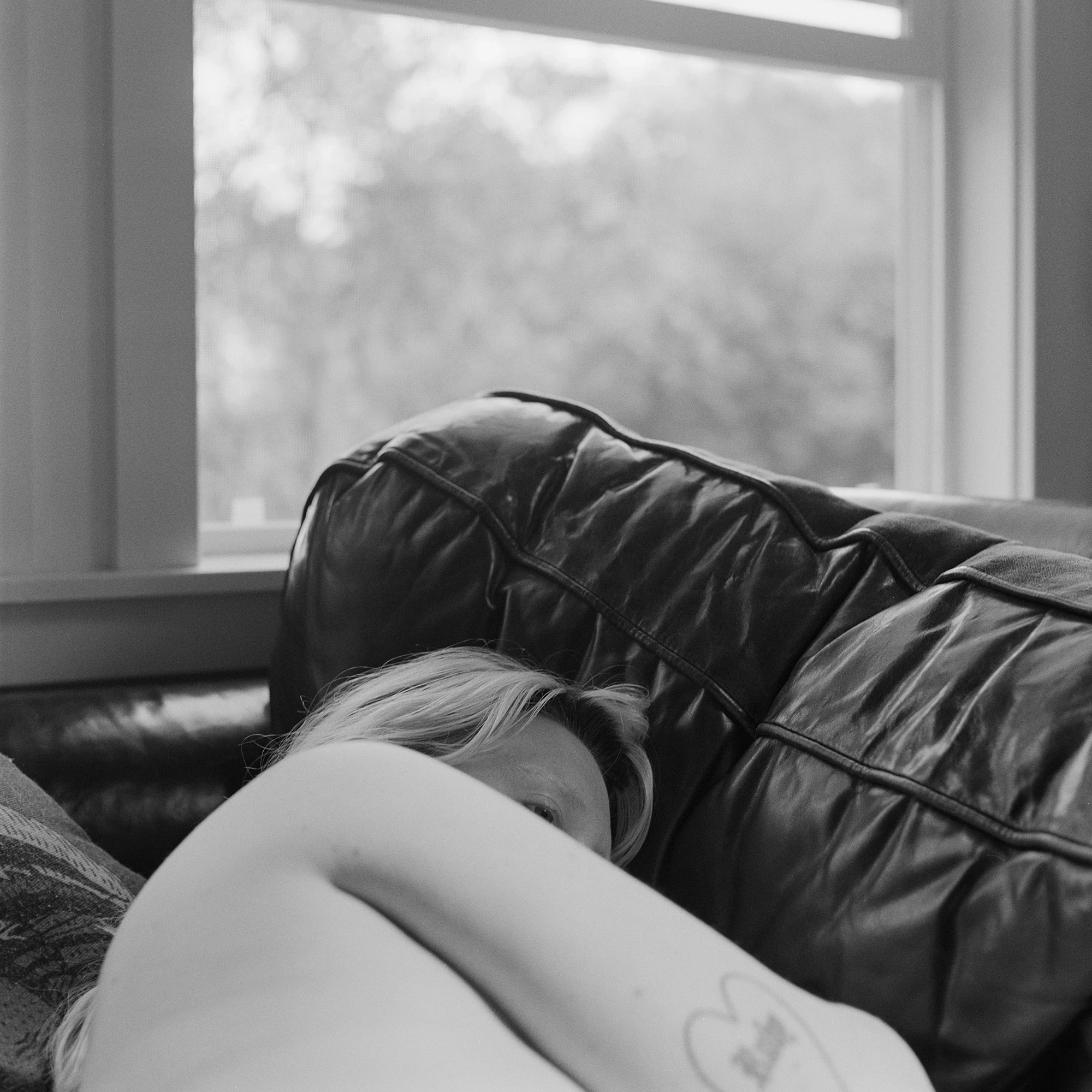 Eternal Return by Benjamin Young, photographed by Benjamin Young — a figure lies half-hidden behind a creased black leather sofa set below a bright bare window.