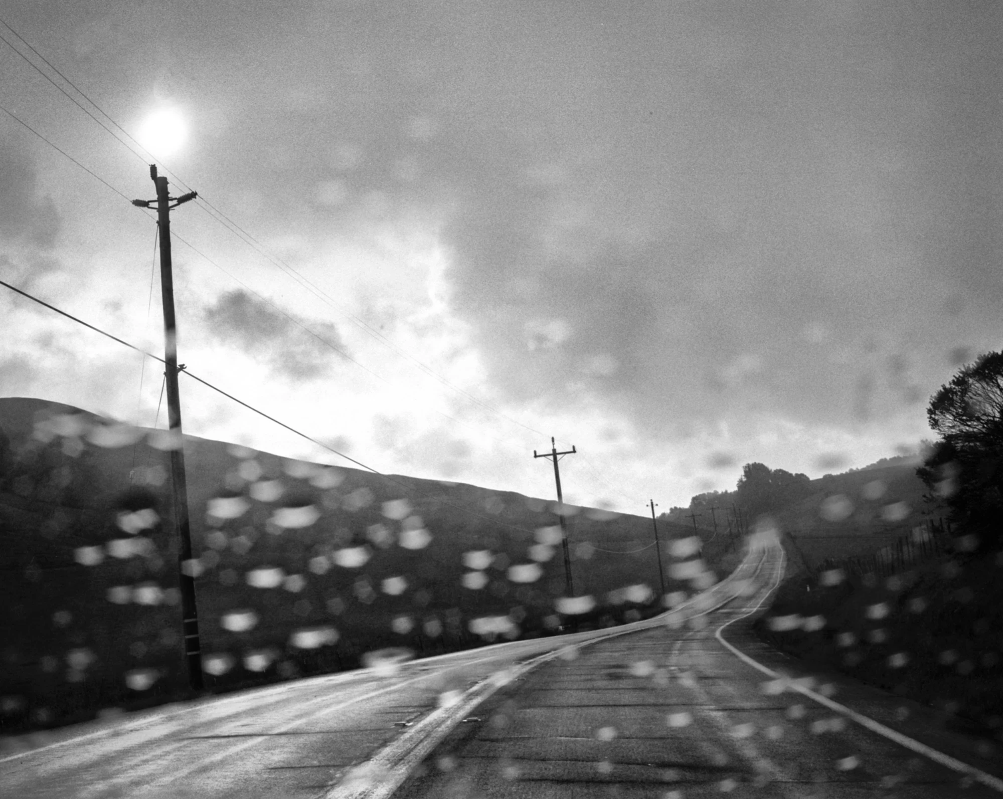 Eternal Return by Benjamin Young, photographed by Benjamin Young — a wet road runs through open hills seen through a rain-flecked windshield, the sun flaring above the ridge.
