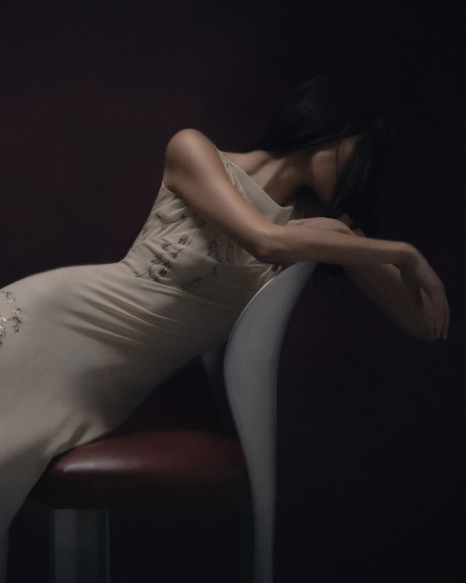 Lilium Chair by Studio NRG — a figure in a pale gown reclines against the curved aluminium backrest above an oxblood leather cushion in low light.