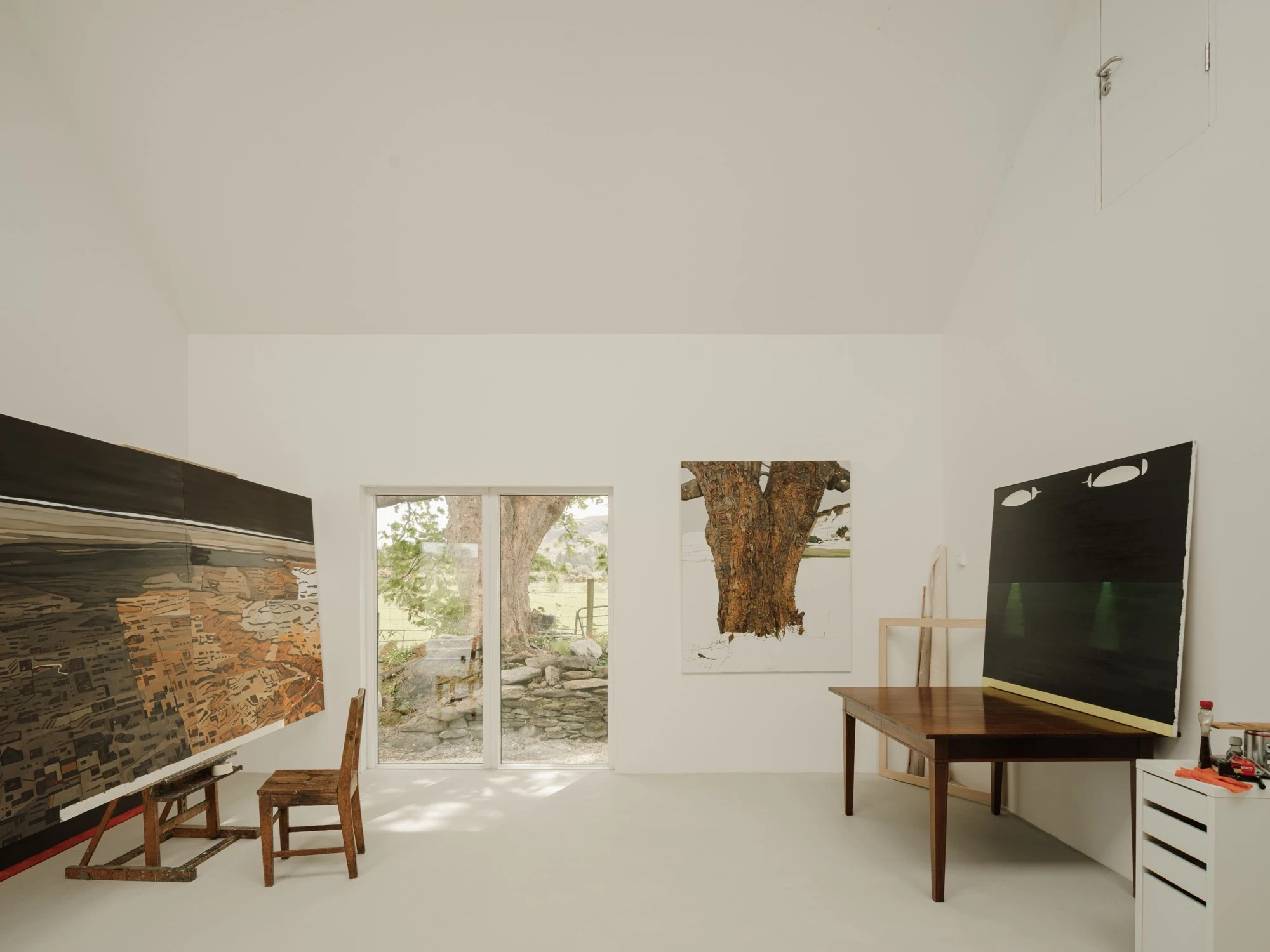 Moss by Pasparakis Friel, photographed by Peter Molloy — a tall white studio holds large paintings, a paint-spattered easel, and a timber table before a garden door framing an old tree.