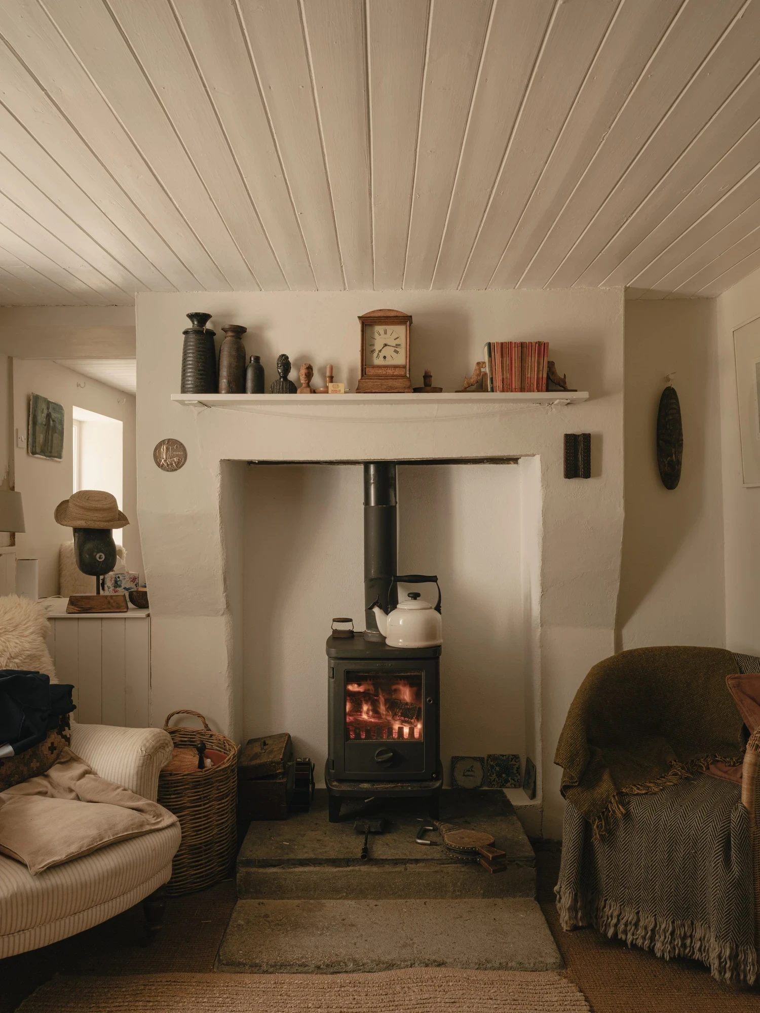 Moss by Pasparakis Friel, photographed by Peter Molloy — a black cast-iron stove burns in a whitewashed cottage hearth beneath a painted tongue-and-groove ceiling and a mantel of ceramics.