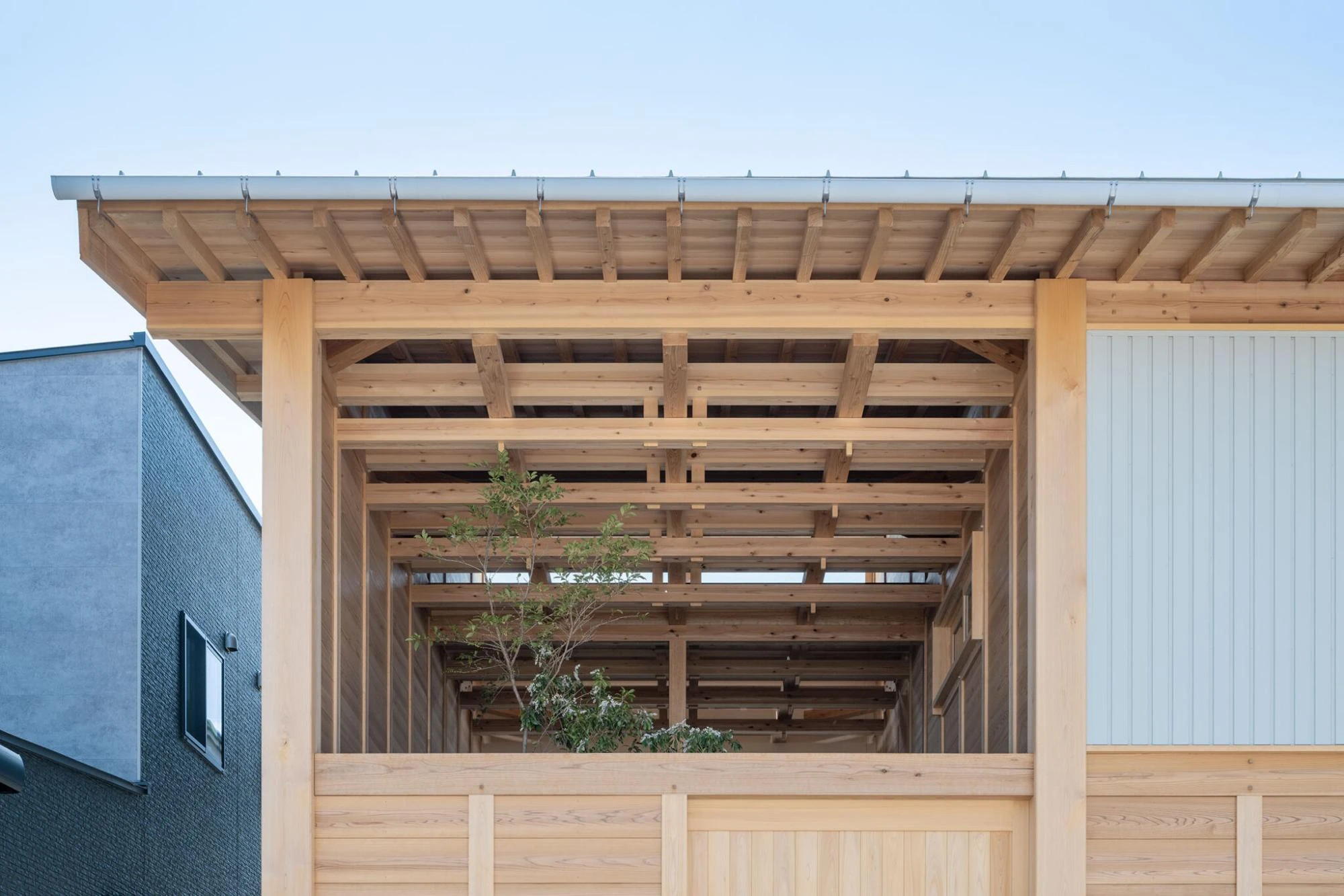 Single Family House in Aichi by Ryota Sekiya, photographed by Shun Fukuda — a close view of the roof eave showing the stacked beam lattice open to the sky, a small tree visible through the timber grid.