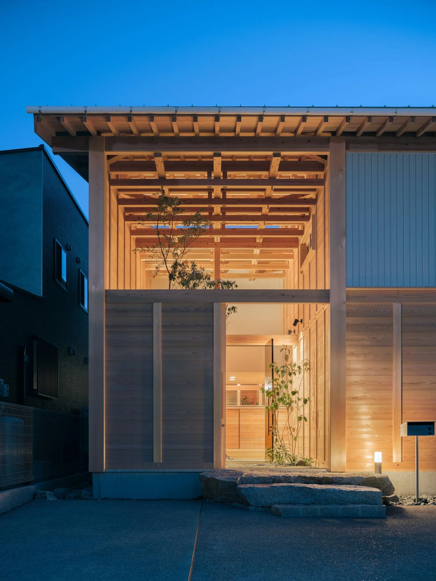 Single Family House in Aichi by Ryota Sekiya, photographed by Shun Fukuda — the entrance void glowing warm at dusk under a deep blue sky, a slim tree lit against pale cedar walls.