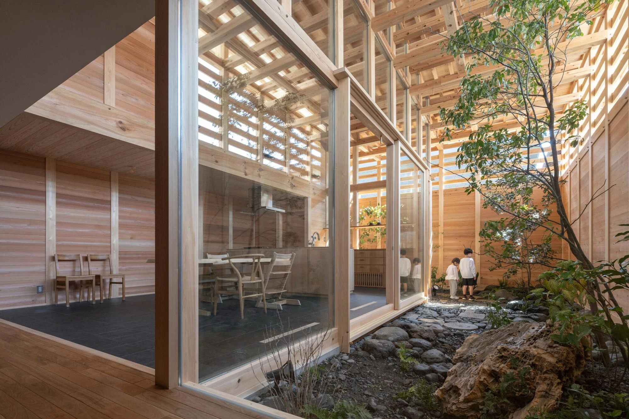 Single Family House in Aichi by Ryota Sekiya, photographed by Shun Fukuda — glazed sliding walls separate the dining room from a stone-and-gravel courtyard where two figures crouch by a tree.