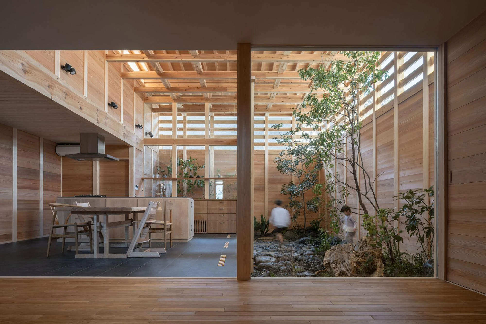 Single Family House in Aichi by Ryota Sekiya, photographed by Shun Fukuda — children move through a courtyard of stones and trees beside the kitchen, seen across a threshold of oak flooring.